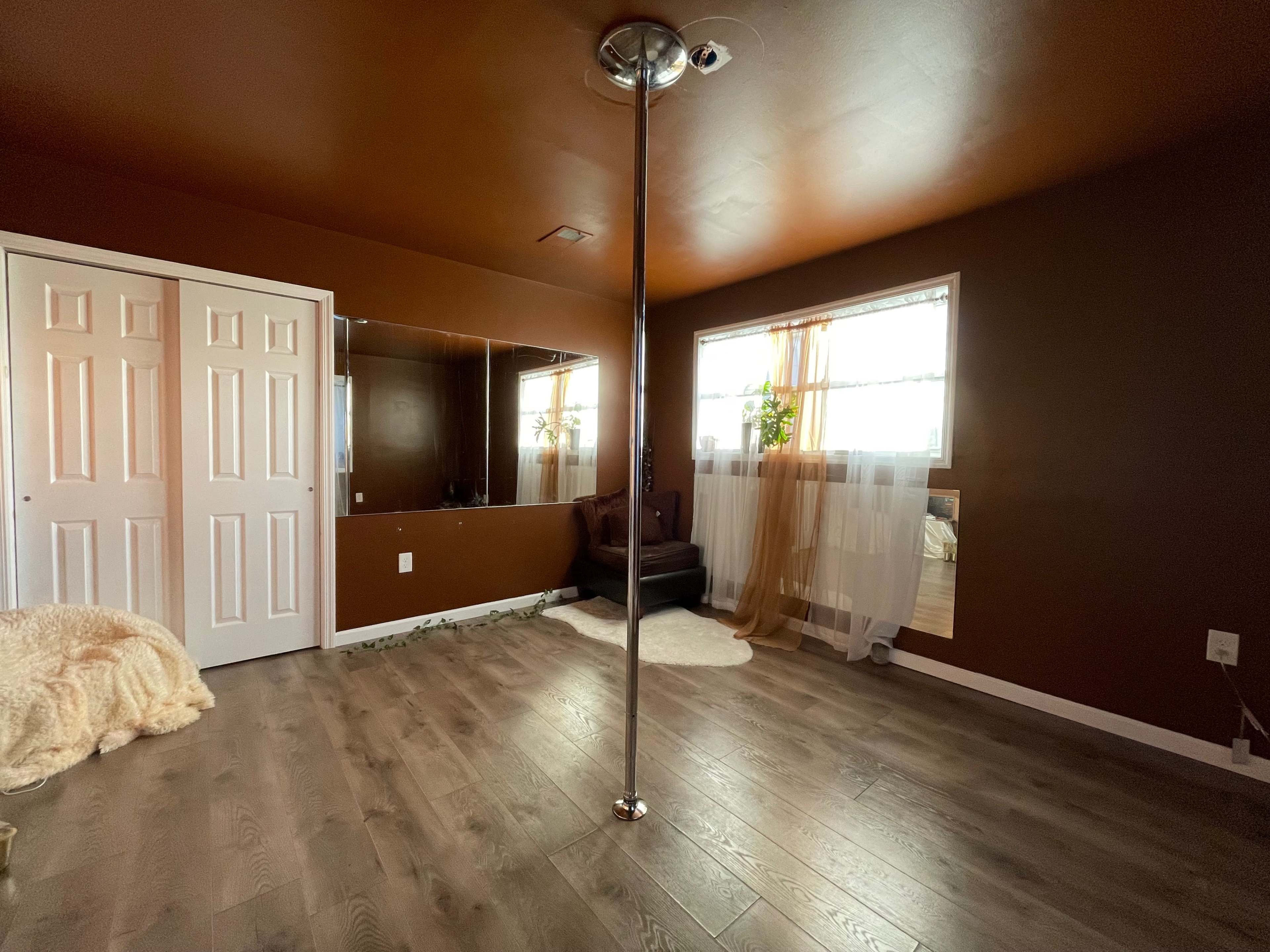 A room with brown walls, a polished wooden floor, a pole in the center, and a mirror on one side.