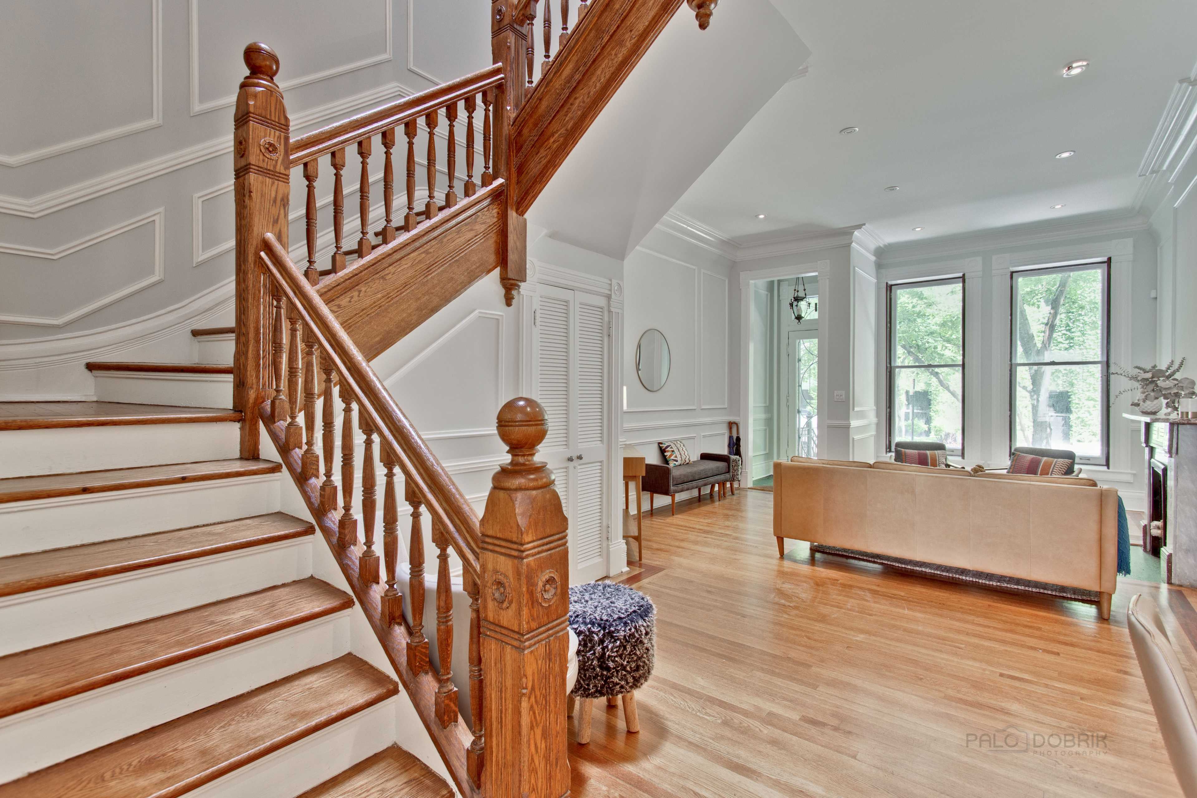 A spacious foyer with a wooden staircase, leading to a living area furnished with a sofa and a chair, all under natural light from large windows.