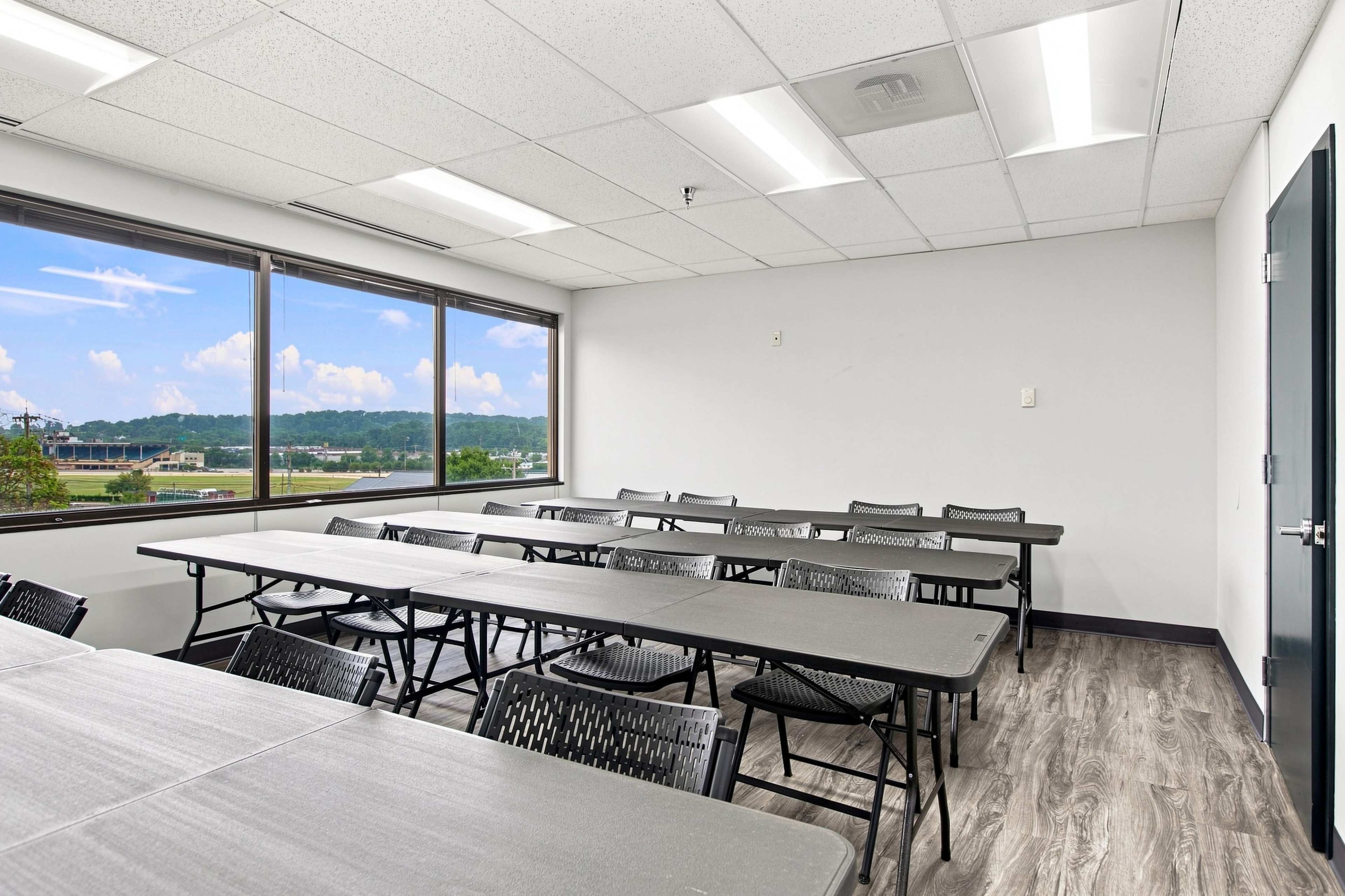 The image shows a spacious, brightly-lit classroom with multiple folding tables arranged in rows and large windows overlooking a landscape.