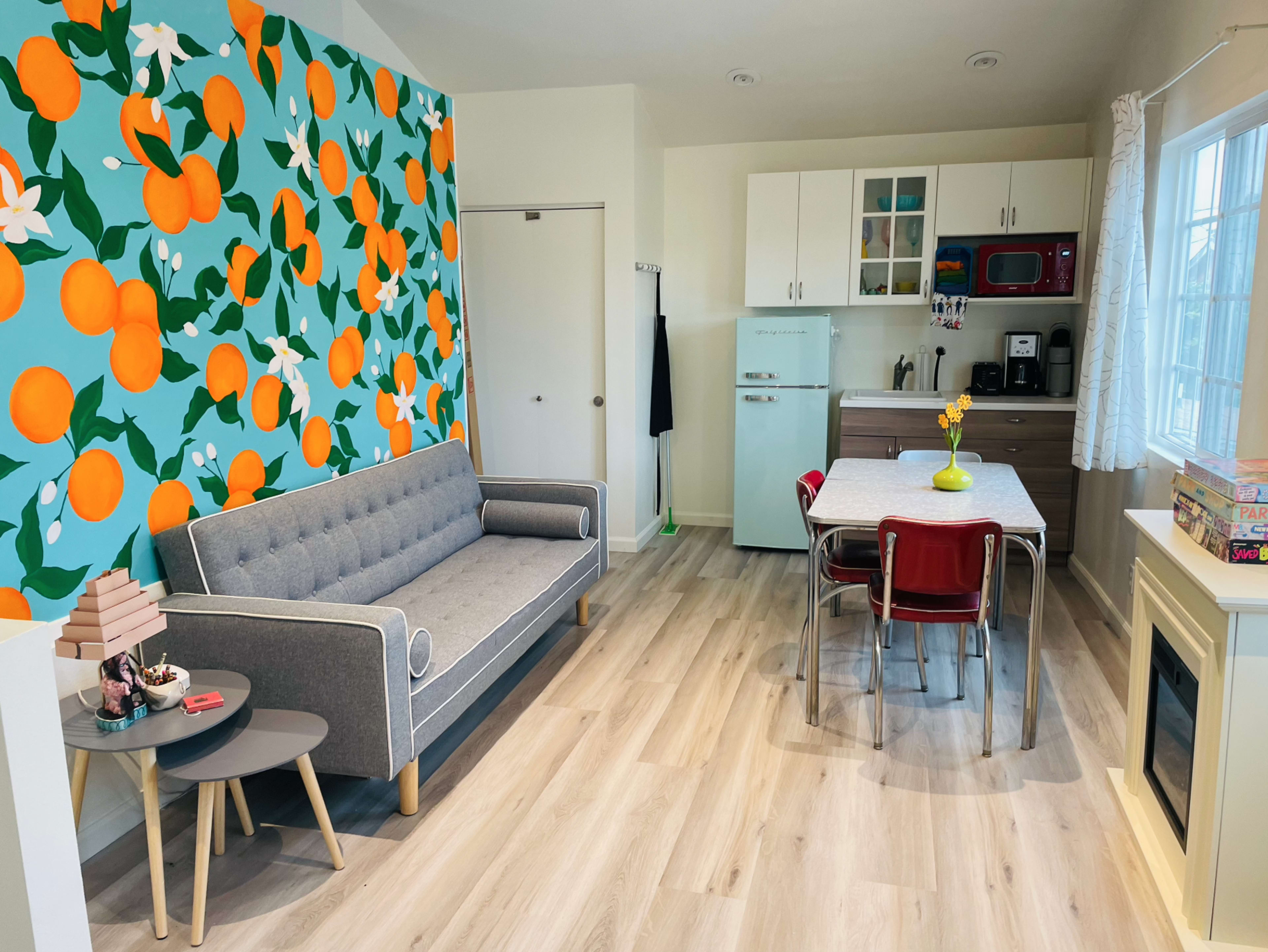 A modern living space with a couch, a dining table with chairs, and a kitchen area, all enhanced by a vibrant orange and floral accent wall.