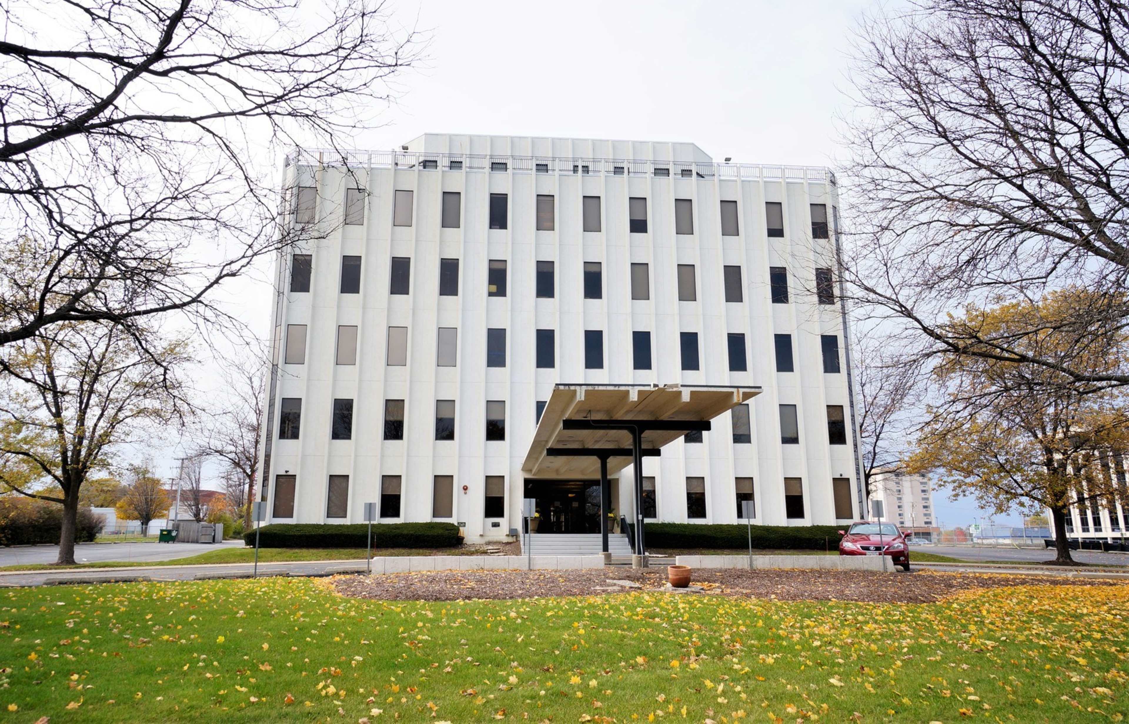 A modern, five-story office building with large windows is situated on a landscaped area with trees and fallen leaves.