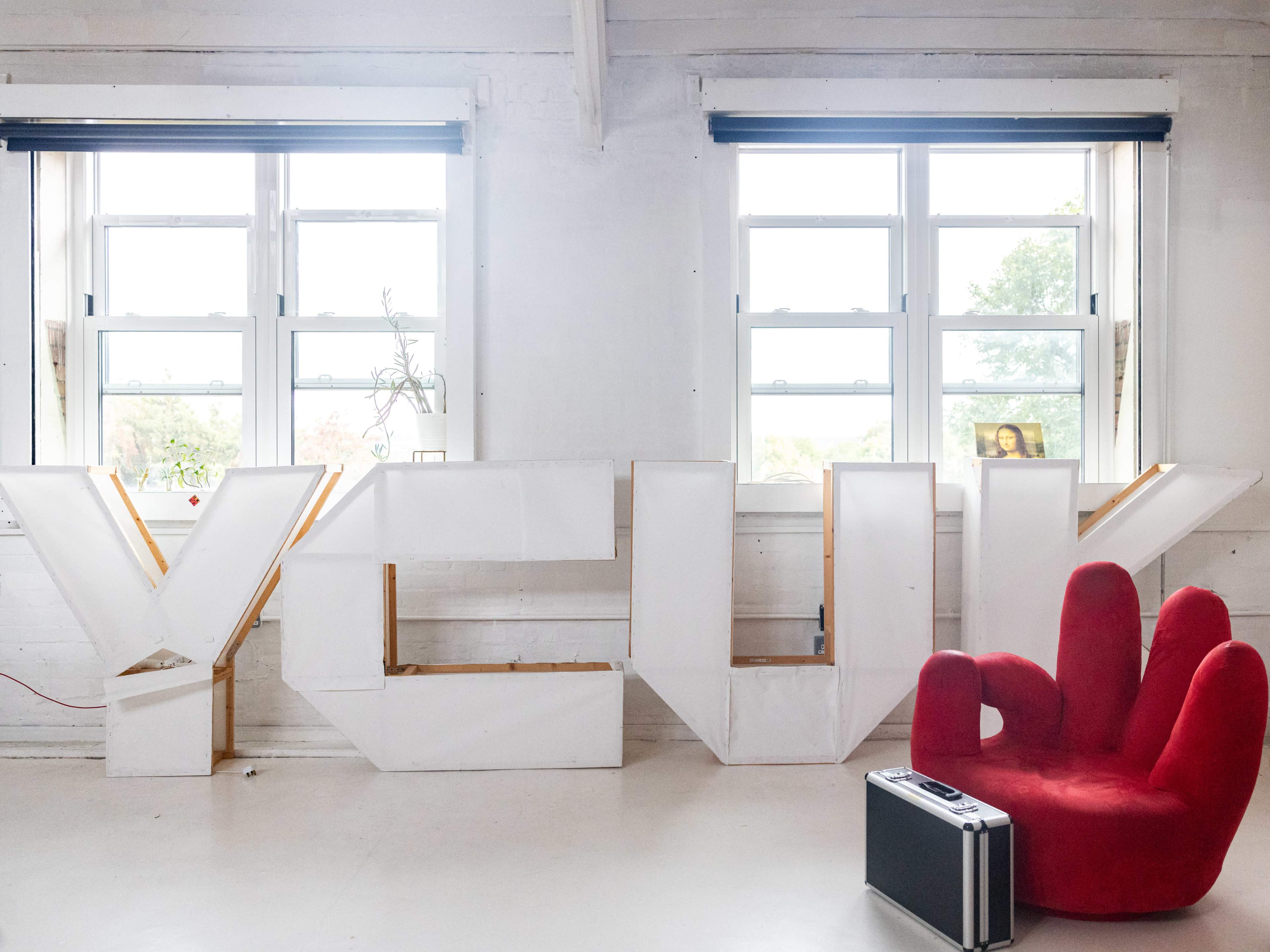 The image features large white letters spelling "YOU" displayed against a wall, with two windows above and a red hand-shaped chair nearby.