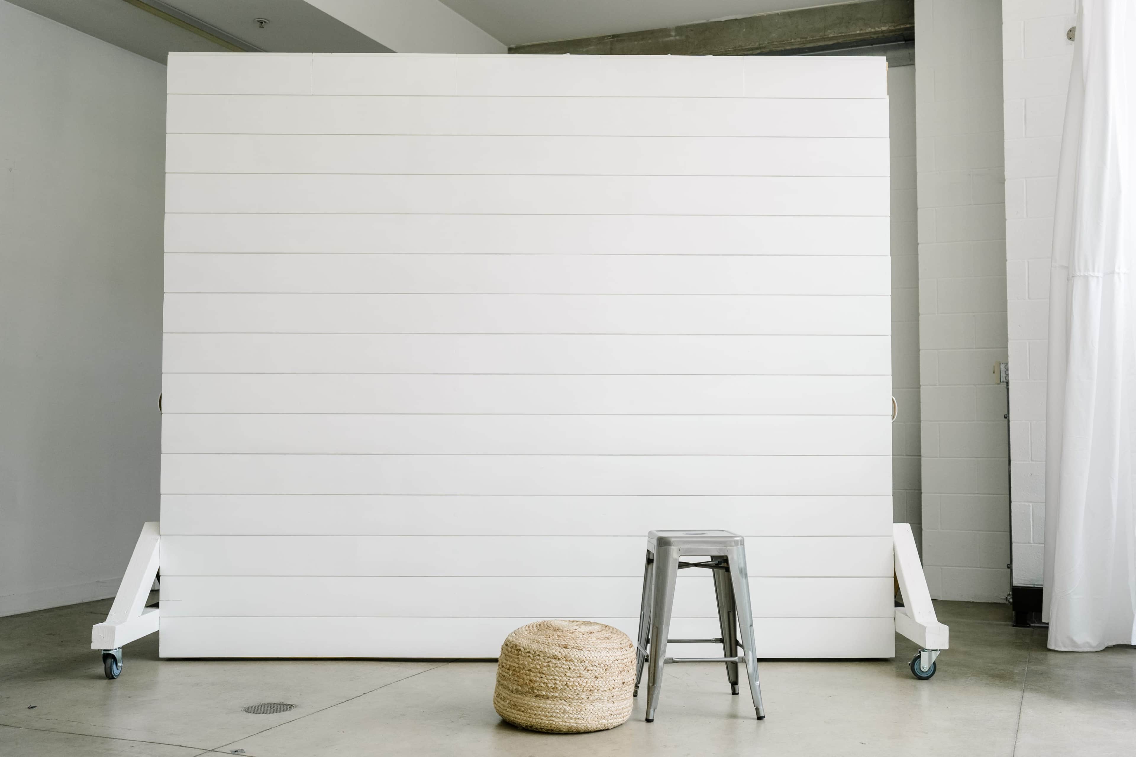 A minimalist studio setup featuring a large white wooden backdrop, a metallic stool, and a woven round pouf on a concrete floor.