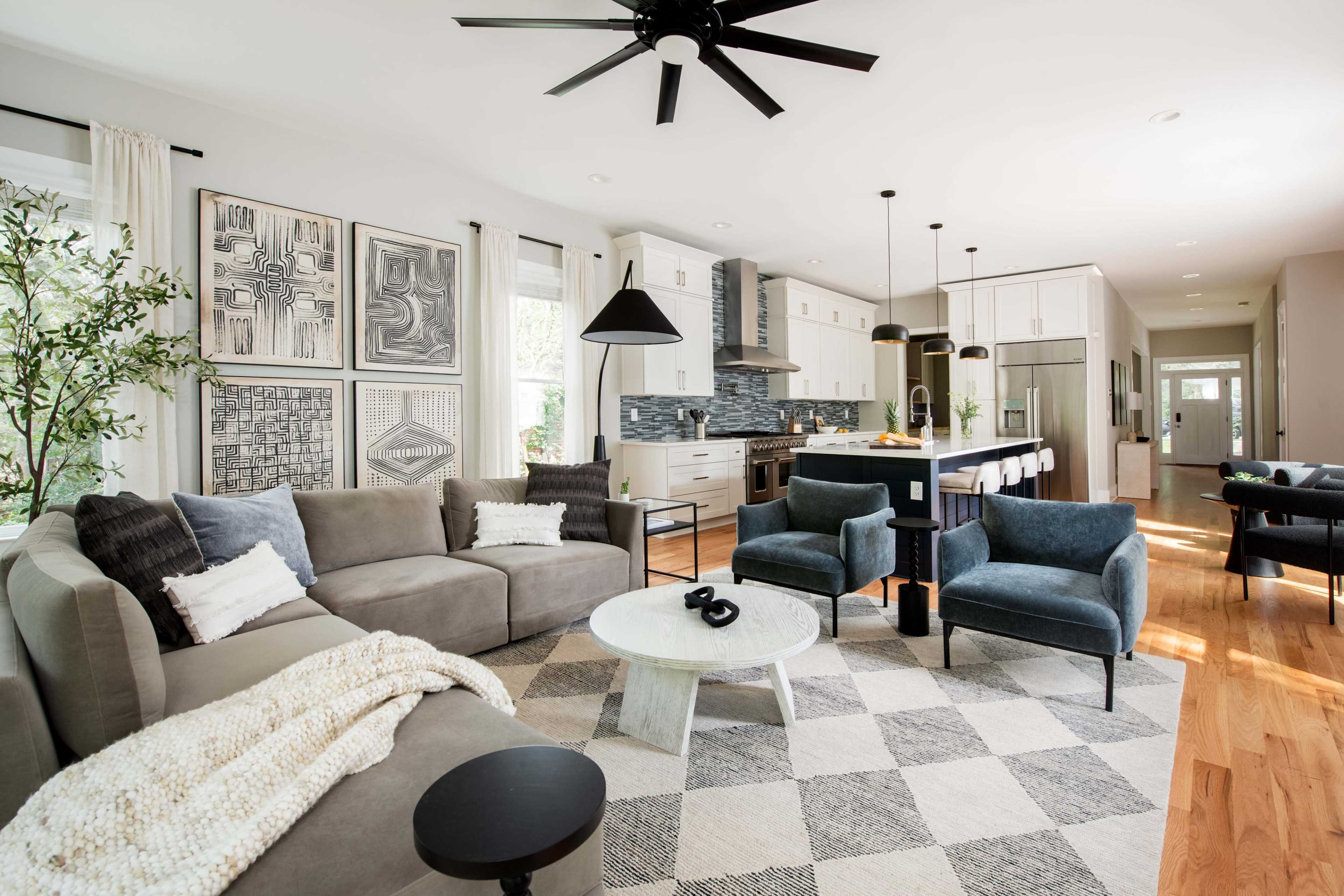 A modern living room features a large gray sectional sofa, a round coffee table, and patterned area rug, with artwork on the walls and a sleek kitchen visible in the background.