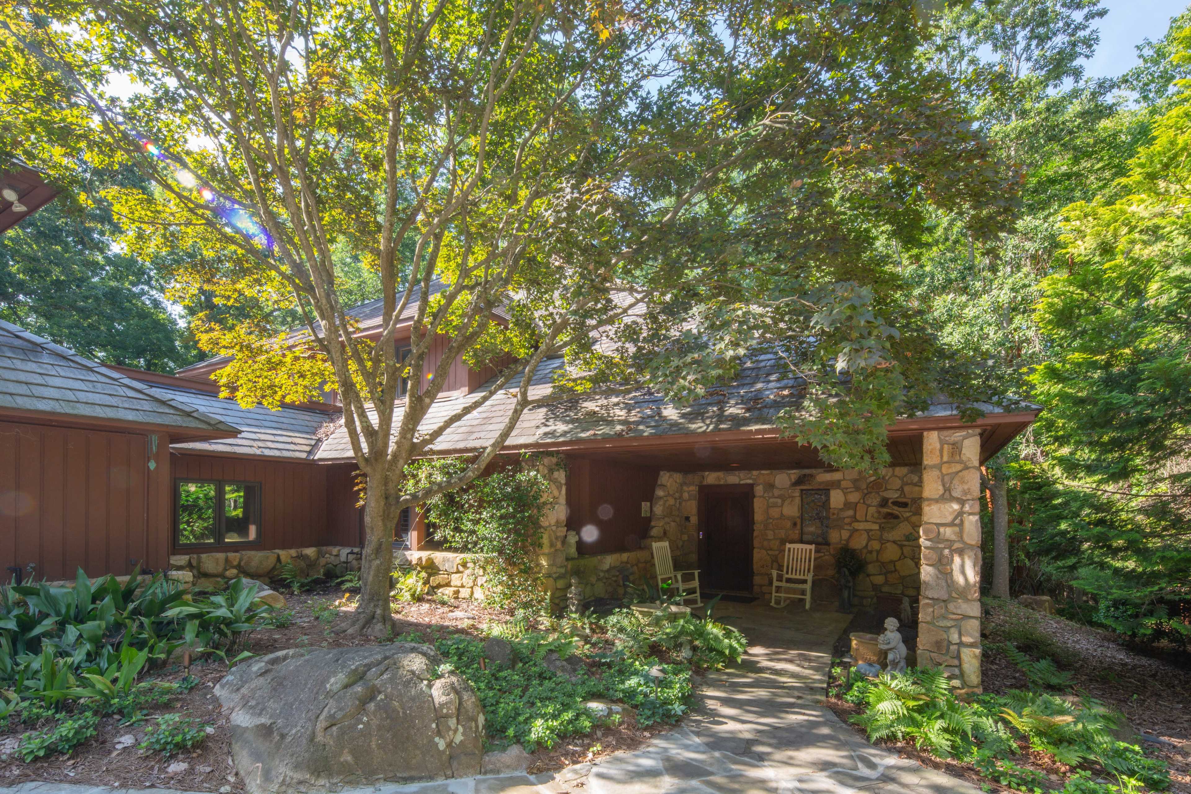 A stone pathway leads to the entrance of a house surrounded by lush greenery and a large tree.