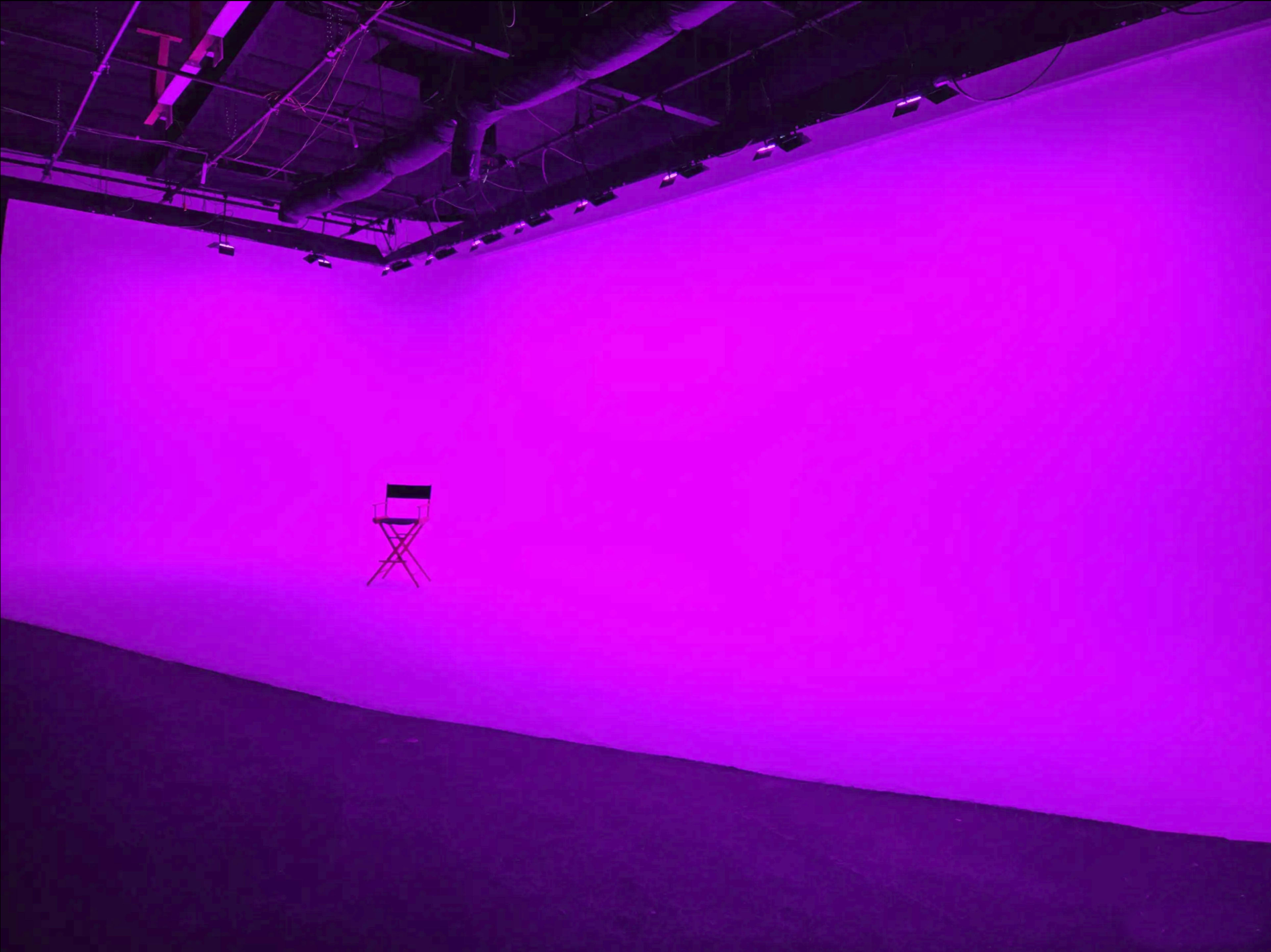 A vacant black director's chair is positioned in front of a solid purple backdrop in a studio space.