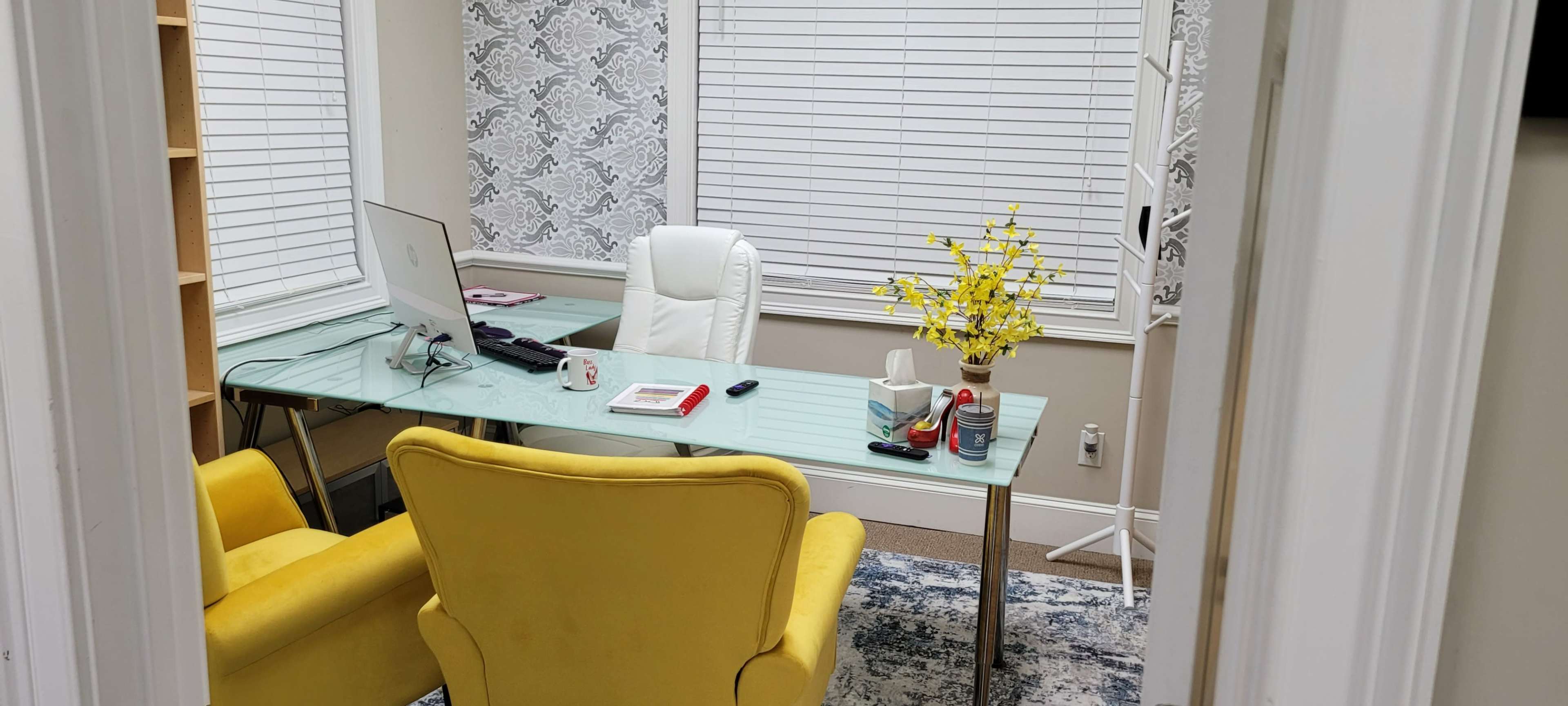 A modern office space features a glass desk with an iMac, two yellow chairs, and a vase of yellow flowers, all situated in a well-lit room with patterned wallpaper.