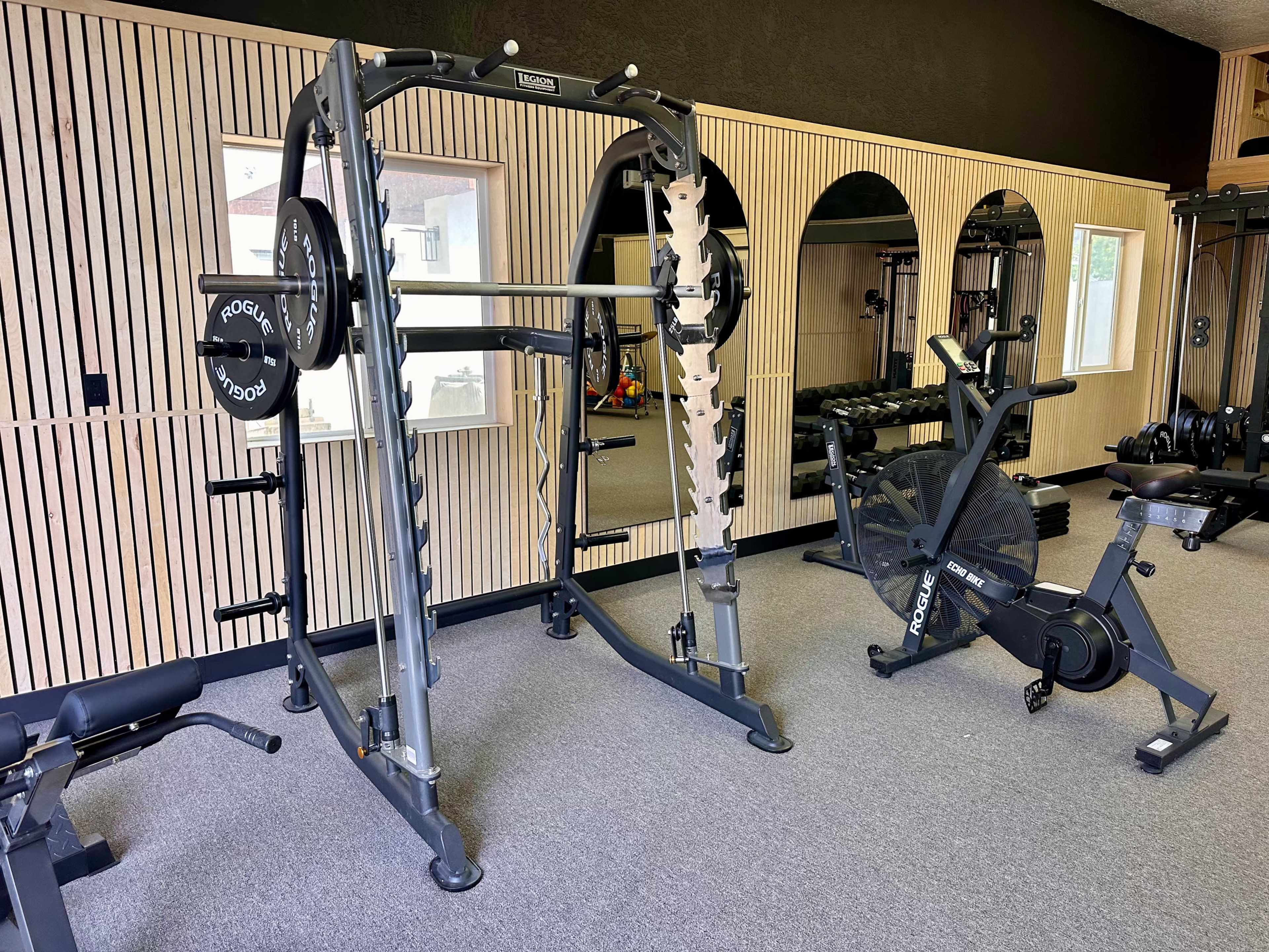 The image shows a modern gym with weightlifting equipment, including a squat rack and an exercise bike, set against wood-paneled walls and large mirrors.