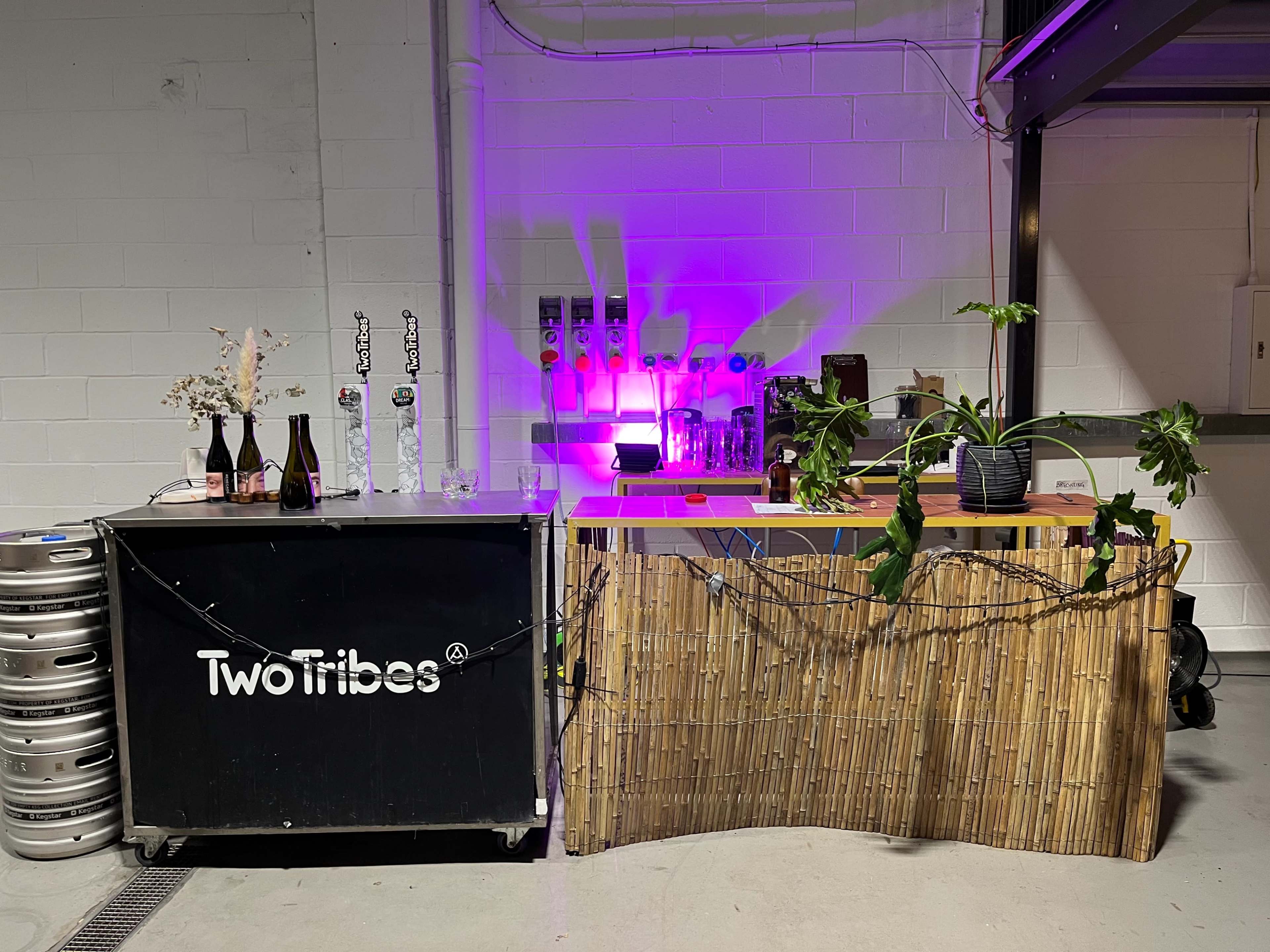A bar area is set up with a black cart labeled "Two Tribes," a bamboo partition, and various bottles on display under purple lighting.