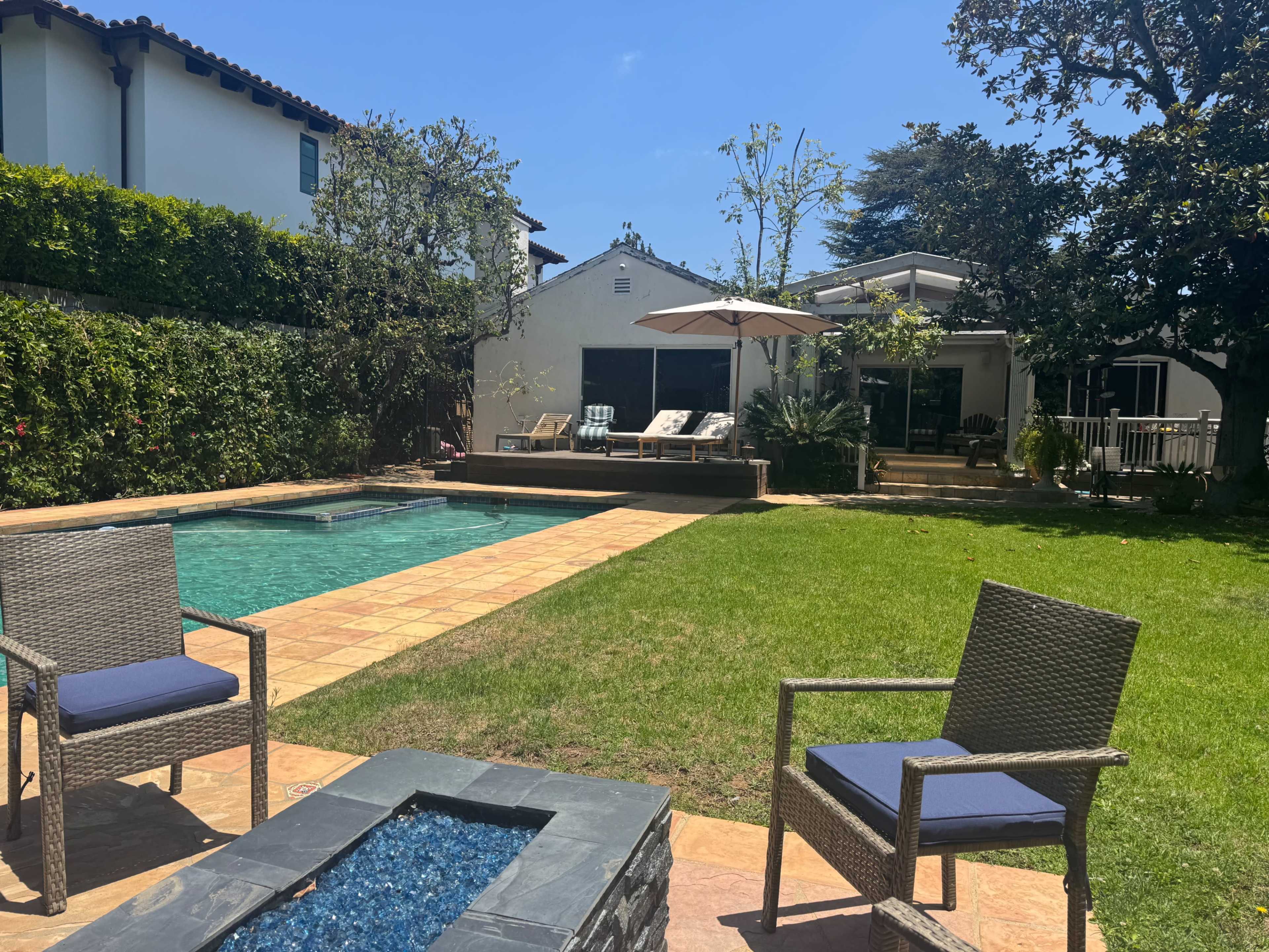 A well-maintained backyard features a swimming pool, lounge chairs, and a fire pit surrounded by green grass and shrubbery.