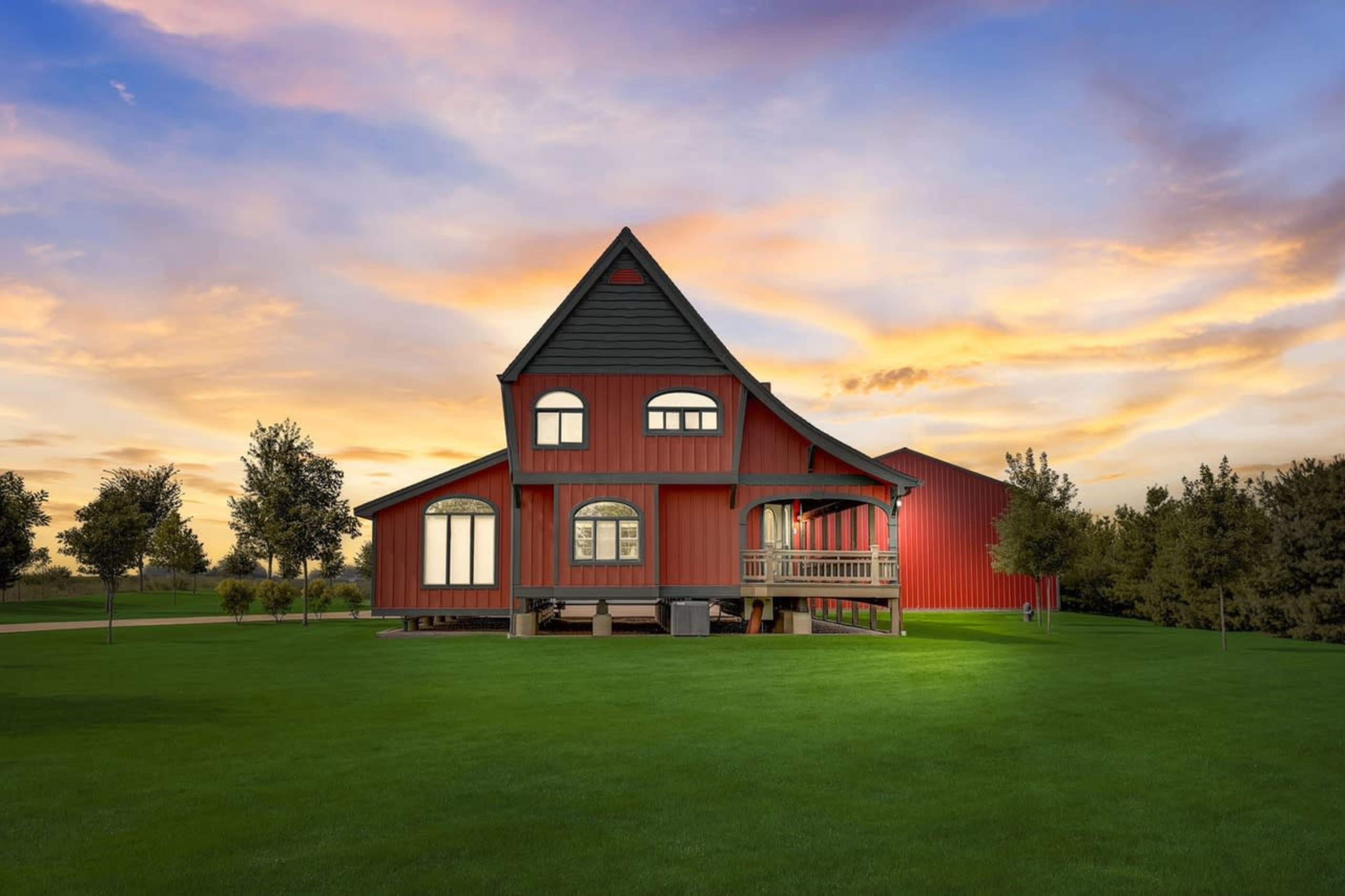 A red farmhouse with a sloped roof and large windows is set on green grass under a colorful sunset sky.