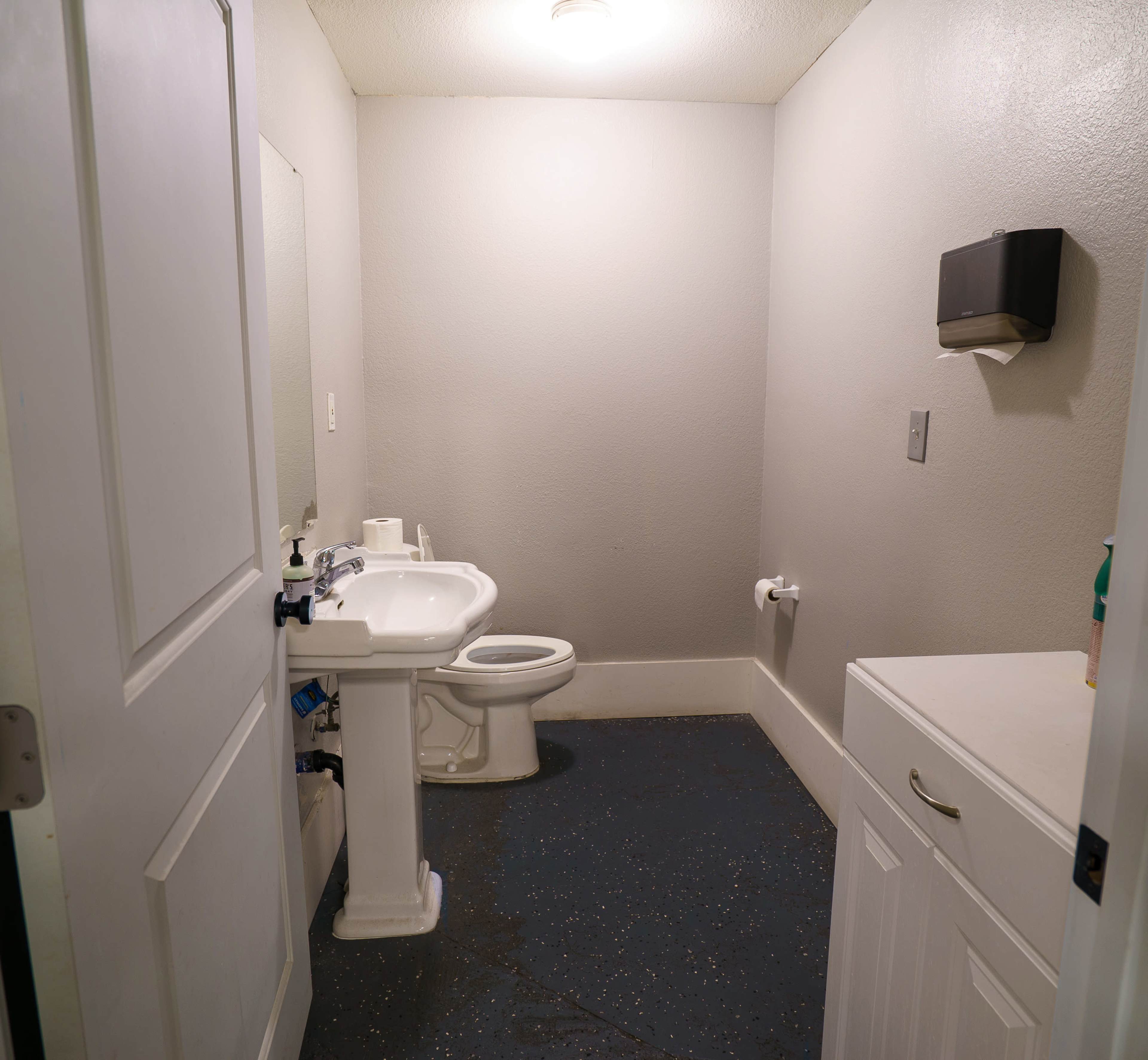 A small, sparsely furnished bathroom features a sink, toilet, and a white cabinet against the wall.