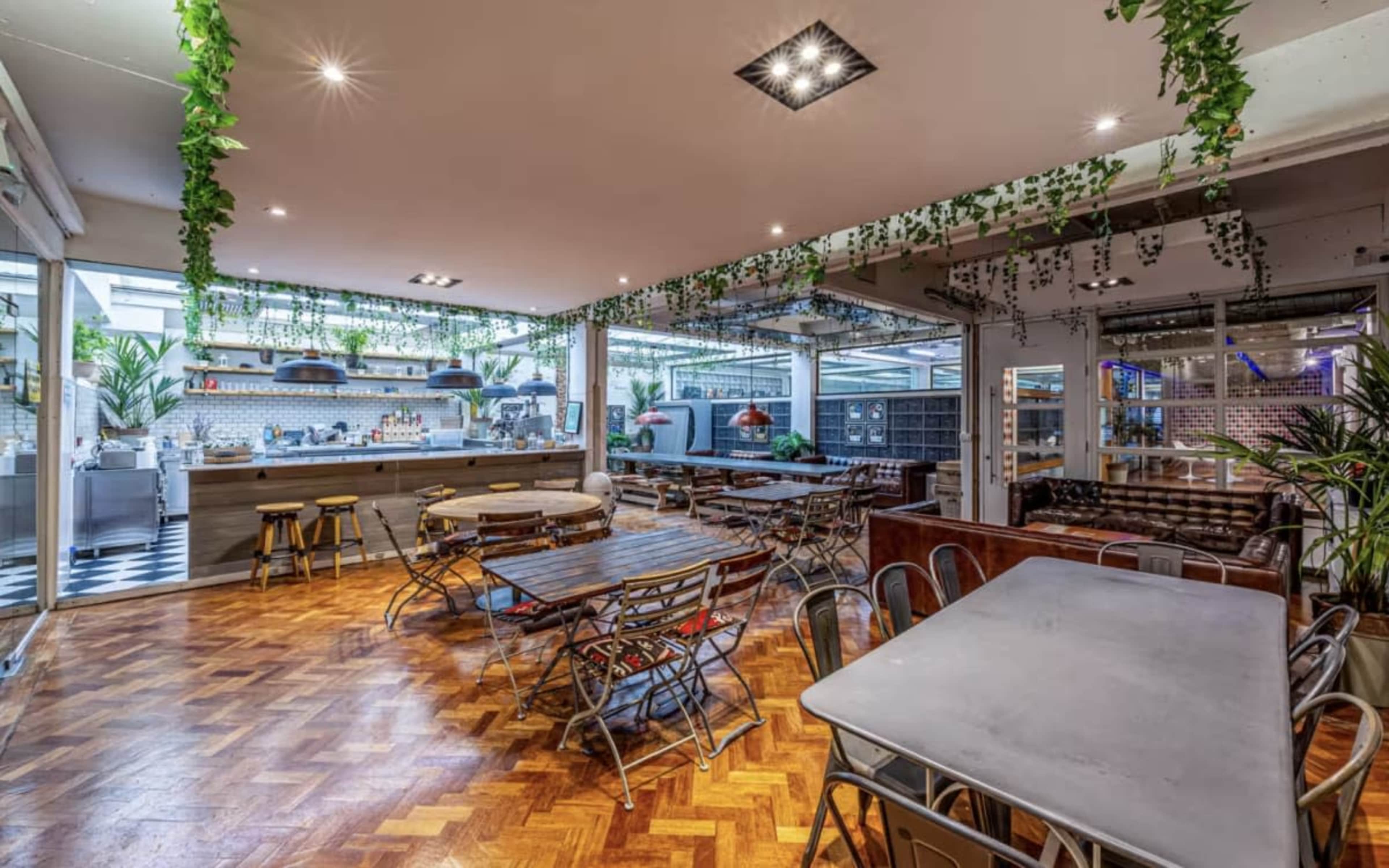 The image shows an indoor dining area featuring wooden floors, metal and wooden tables, and plants hanging from the ceiling, with a bar and kitchen visible in the background.