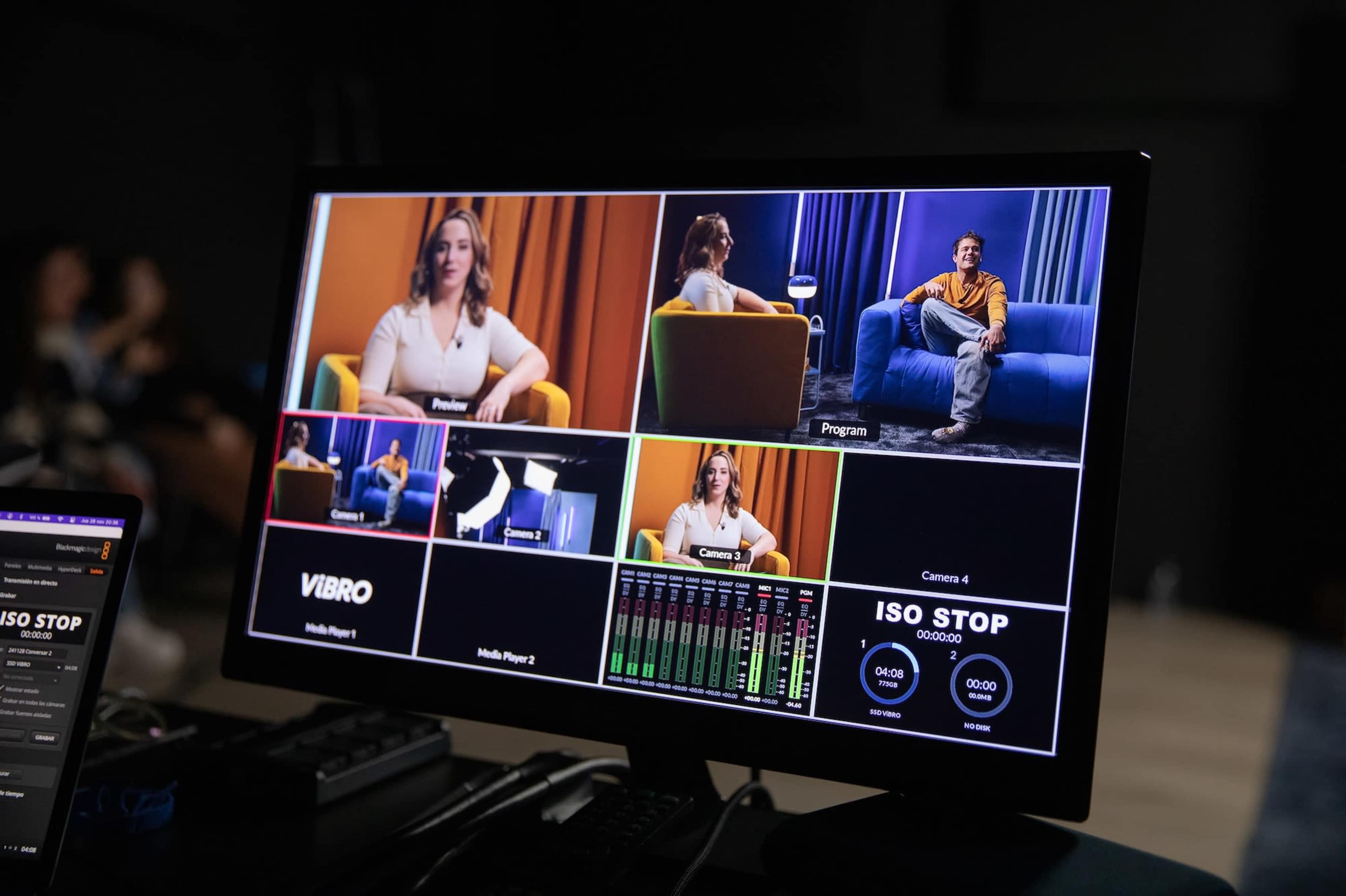 A computer monitor displays a multi-camera setup with video feeds from different angles featuring two people in a studio setting.