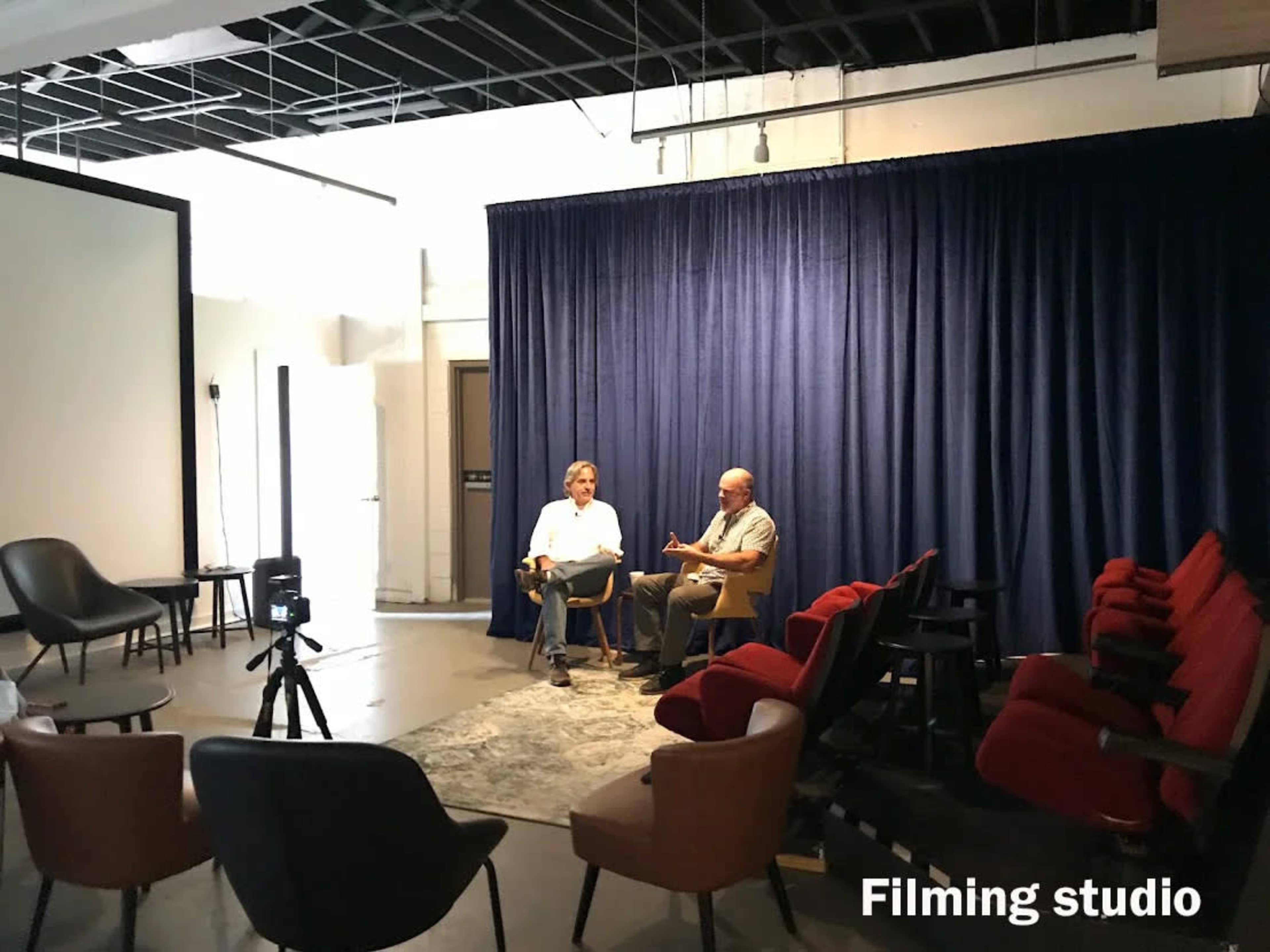 The image shows two men seated in a filming studio with a dark blue curtain backdrop and various chairs arranged nearby.