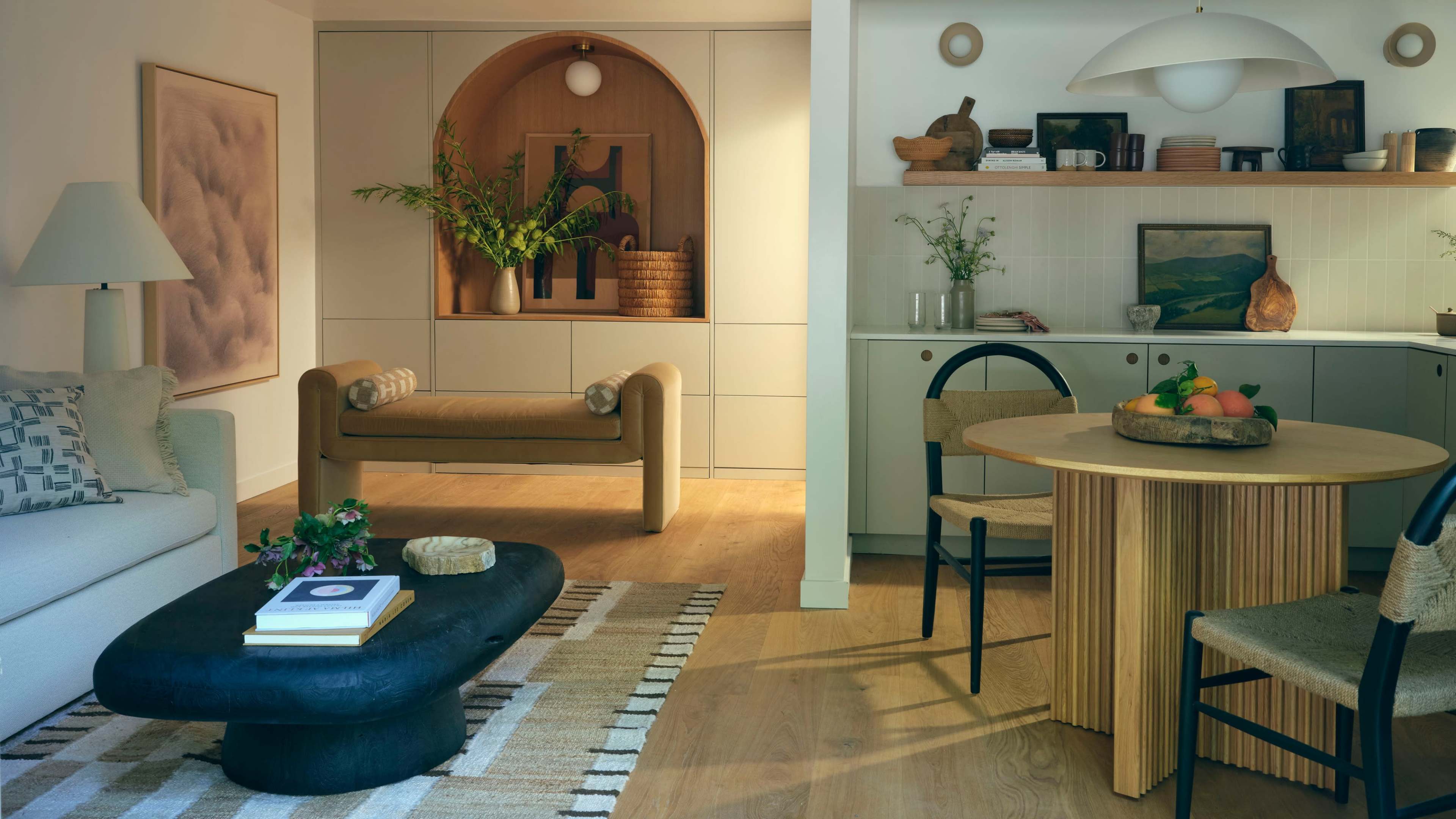 The image shows a modern living space featuring a beige sofa, a rounded wooden bench, a circular dining table, and a kitchen area with light wood accents and decorative plants.