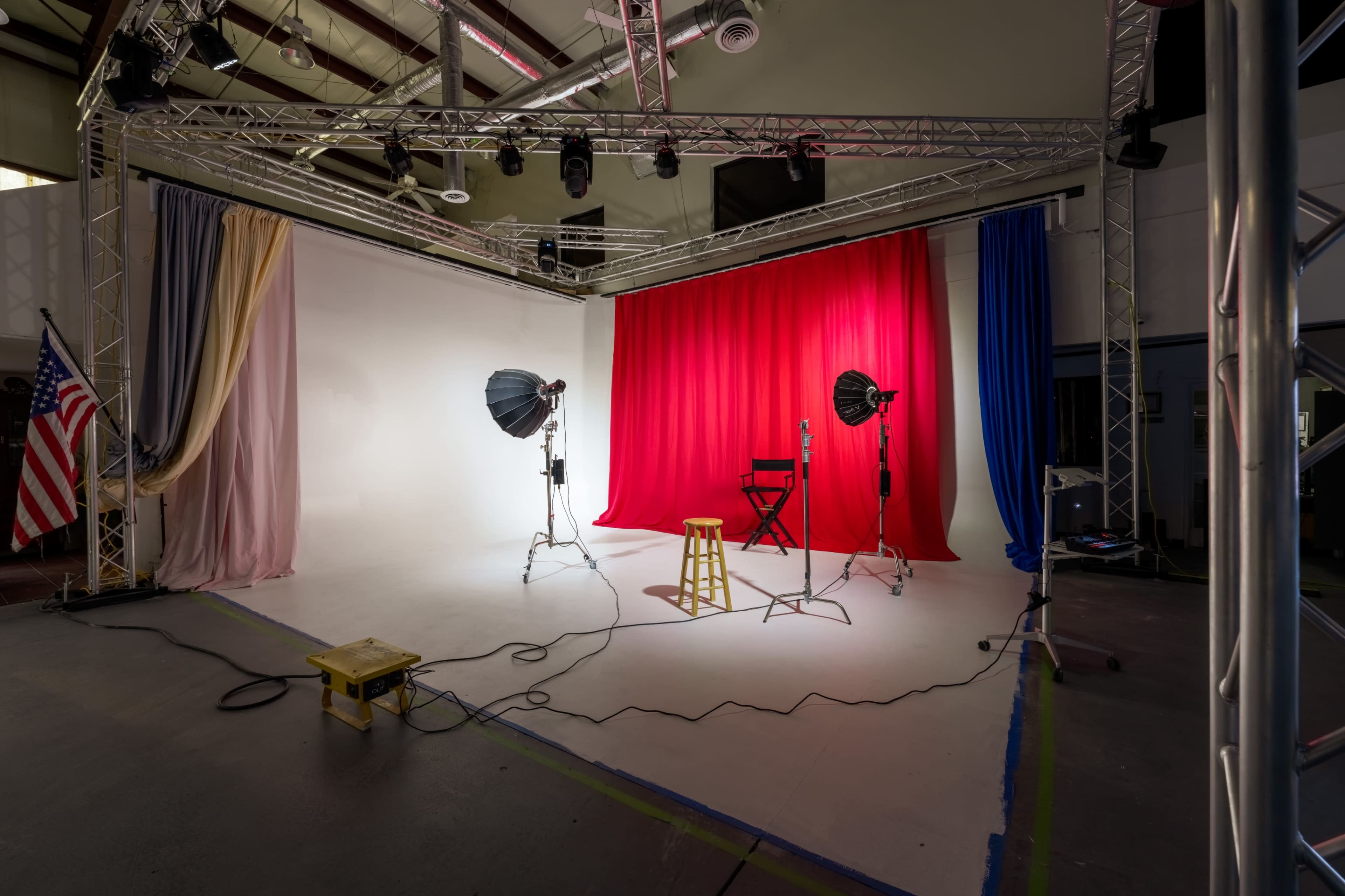The image shows a photography studio setup with a red backdrop, softbox lights, a stool, and an American flag displayed in the corner.