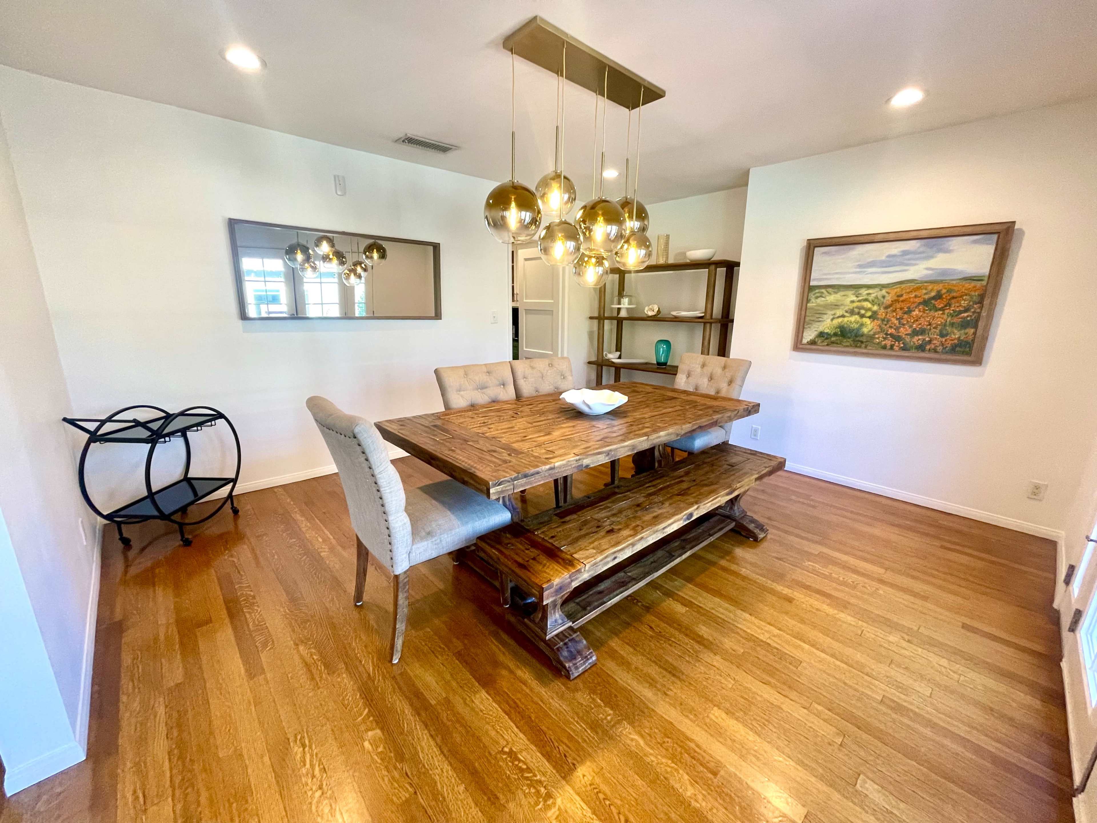 A spacious dining area features a wooden table with a bench and upholstered chairs, illuminated by modern pendant lights, with a decorative mirror and artwork on the walls.
