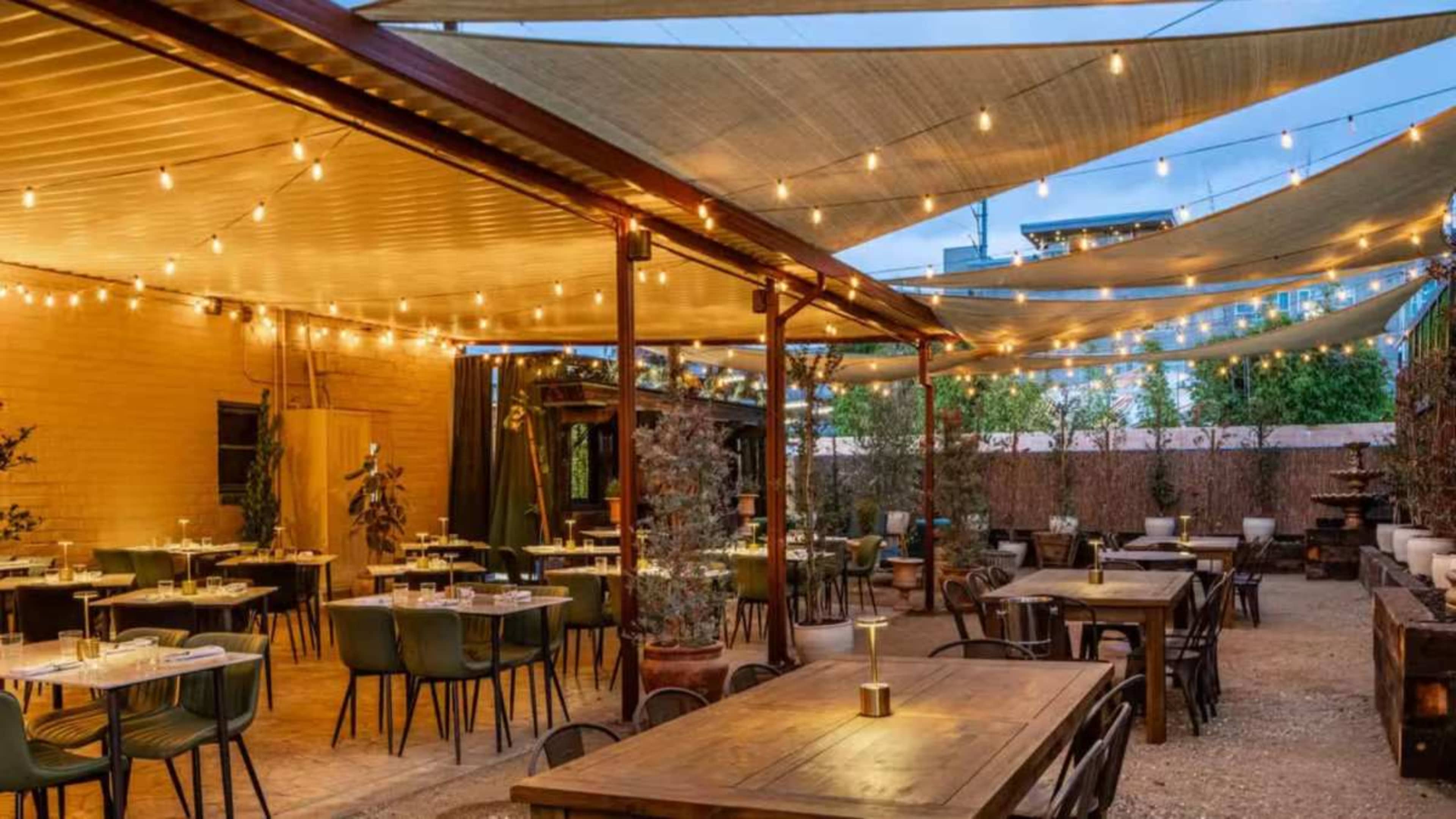 The image shows an outdoor dining area with string lights and shaded tables arranged around a central space.