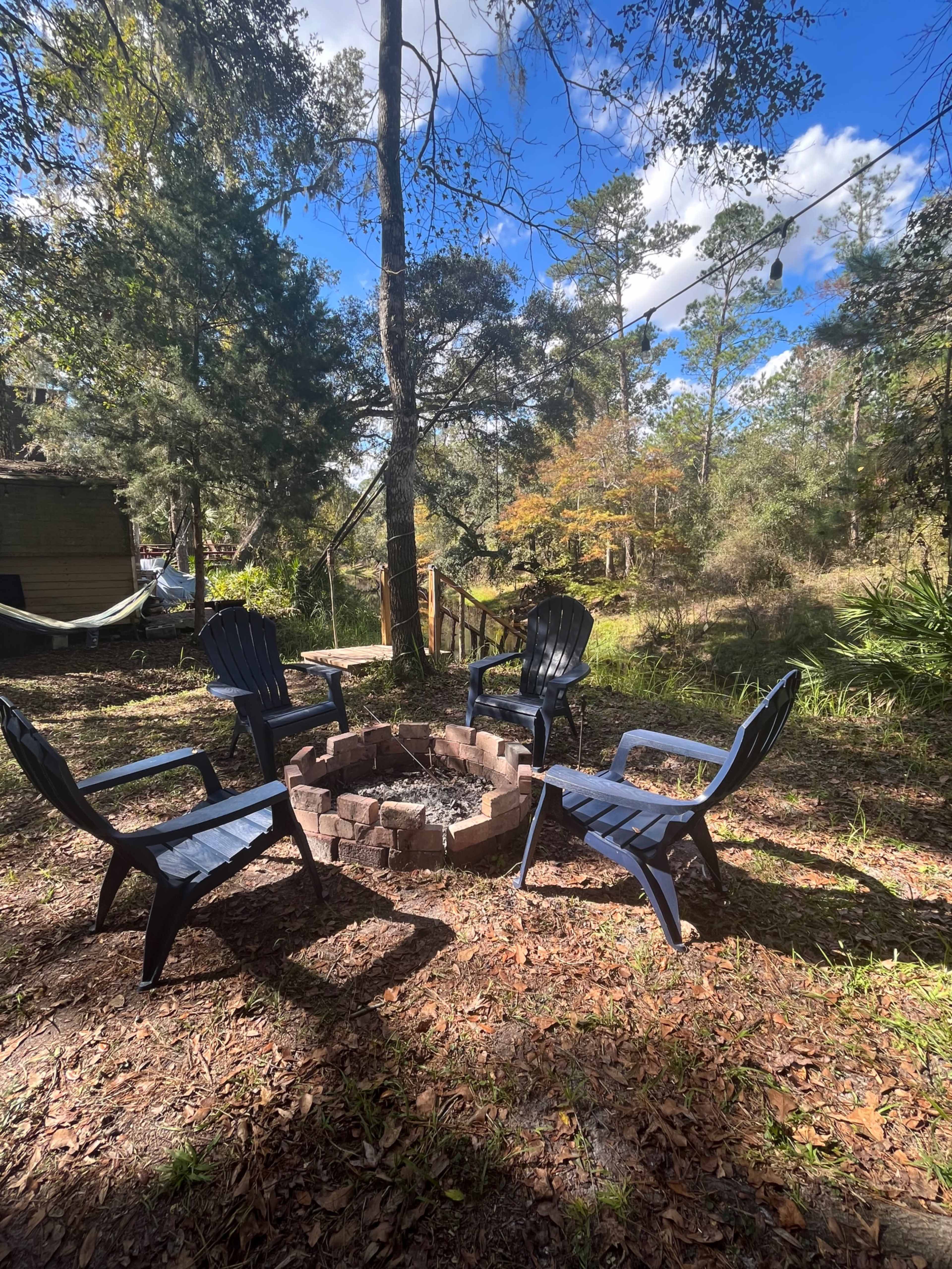 A circular stone fire pit is surrounded by four black plastic chairs in a wooded outdoor setting.