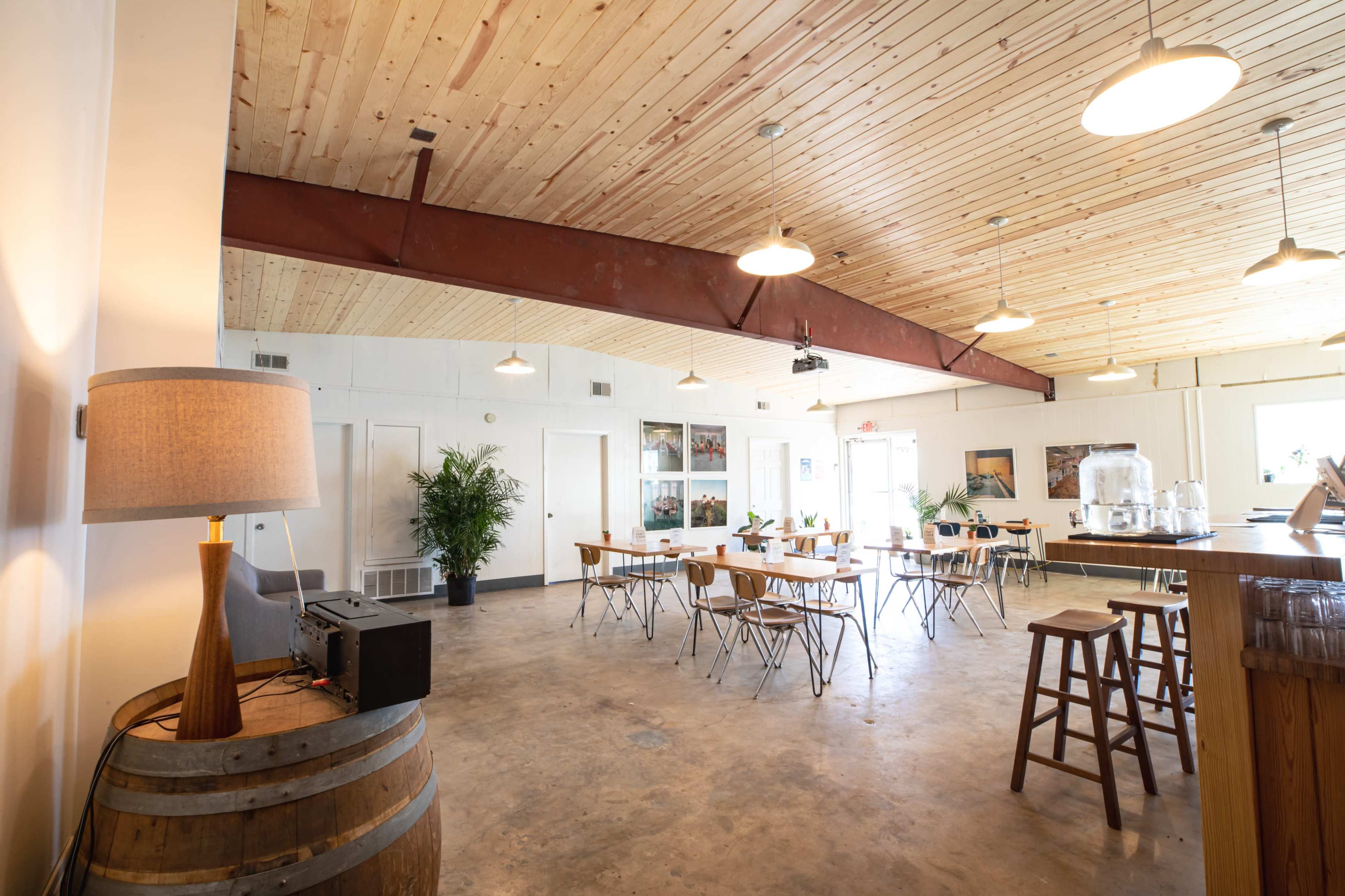 The image shows a spacious room with wooden ceilings, a few tables and chairs arranged, a lamp on a barrel, and large windows letting in natural light.