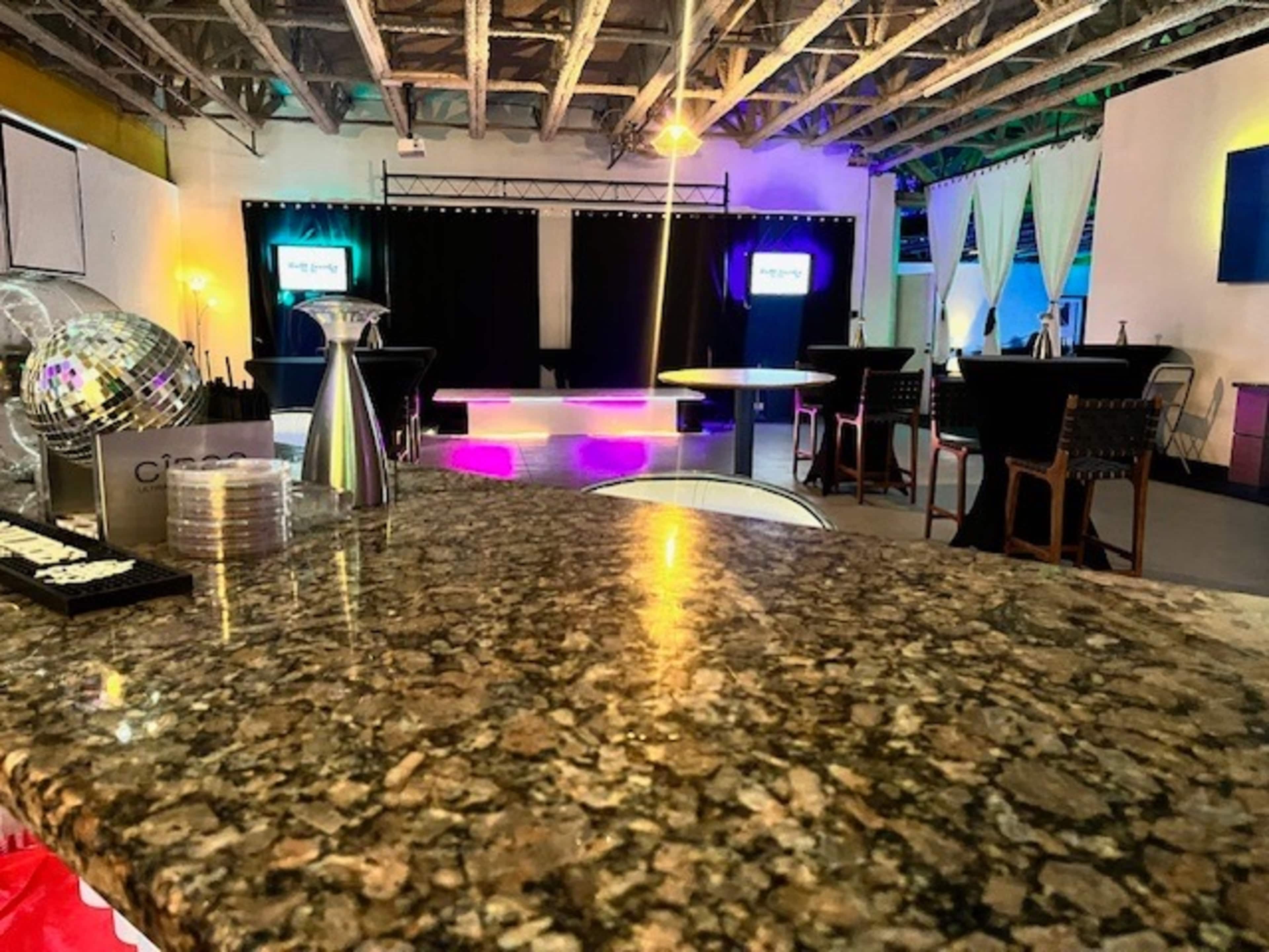 A spacious event venue with a polished granite bar in the foreground, a stage with lights in the background, and several tables and chairs arranged throughout.