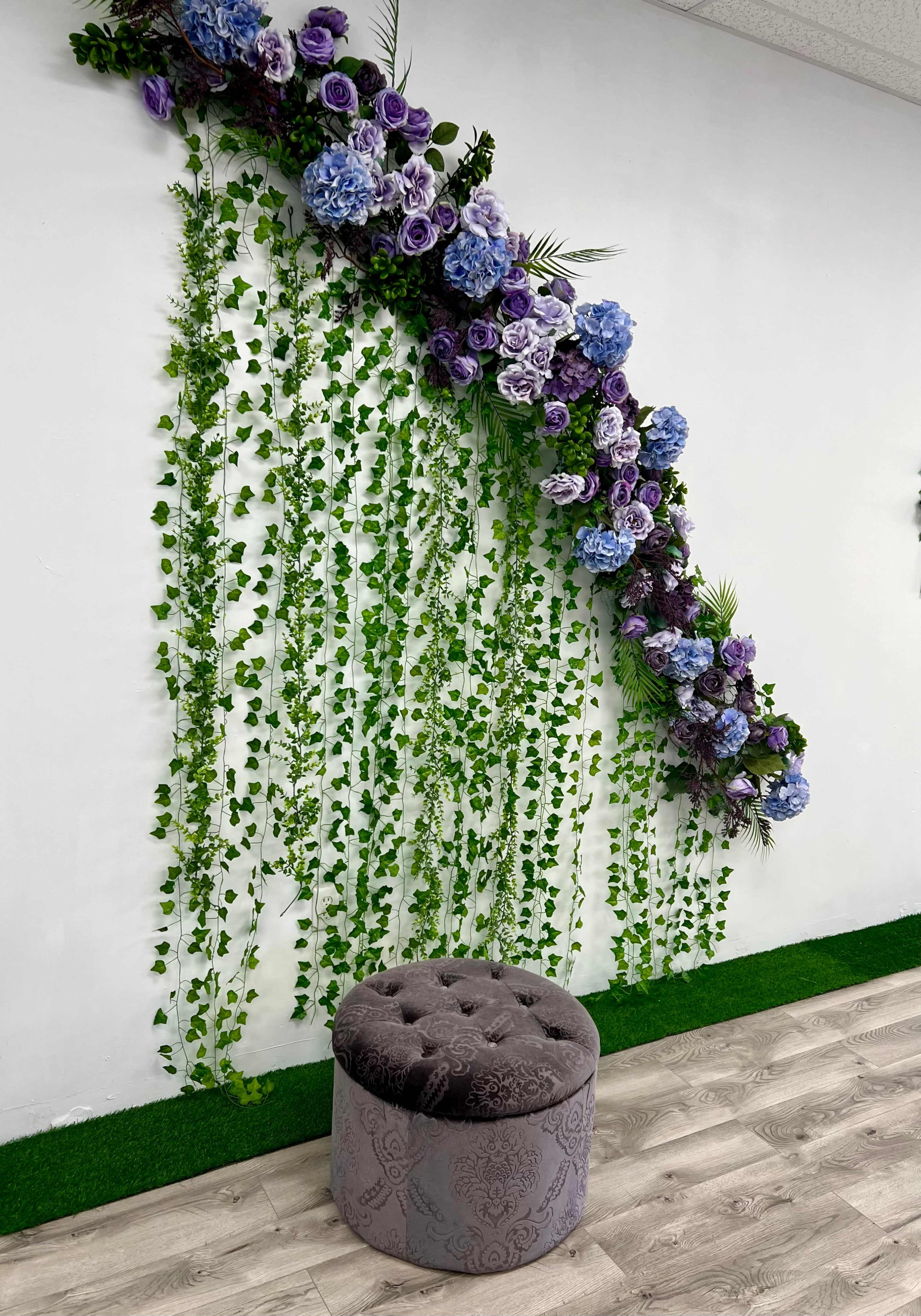 A tufted gray stool sits in front of a wall adorned with cascading greenery and clusters of purple and blue floral arrangements.