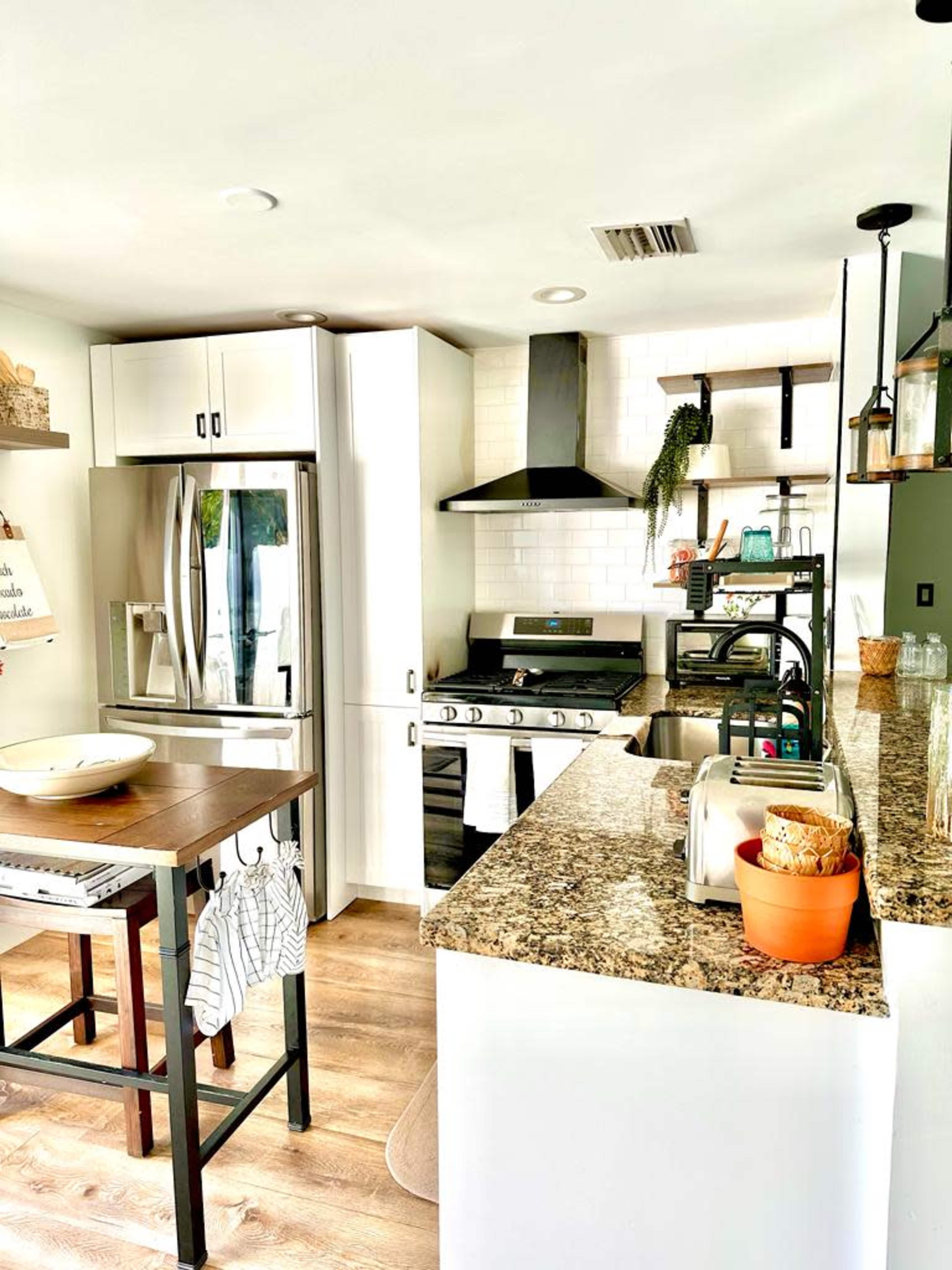 The image shows a modern kitchen with white cabinets, a stainless steel refrigerator, a stove, and a wooden dining table.
