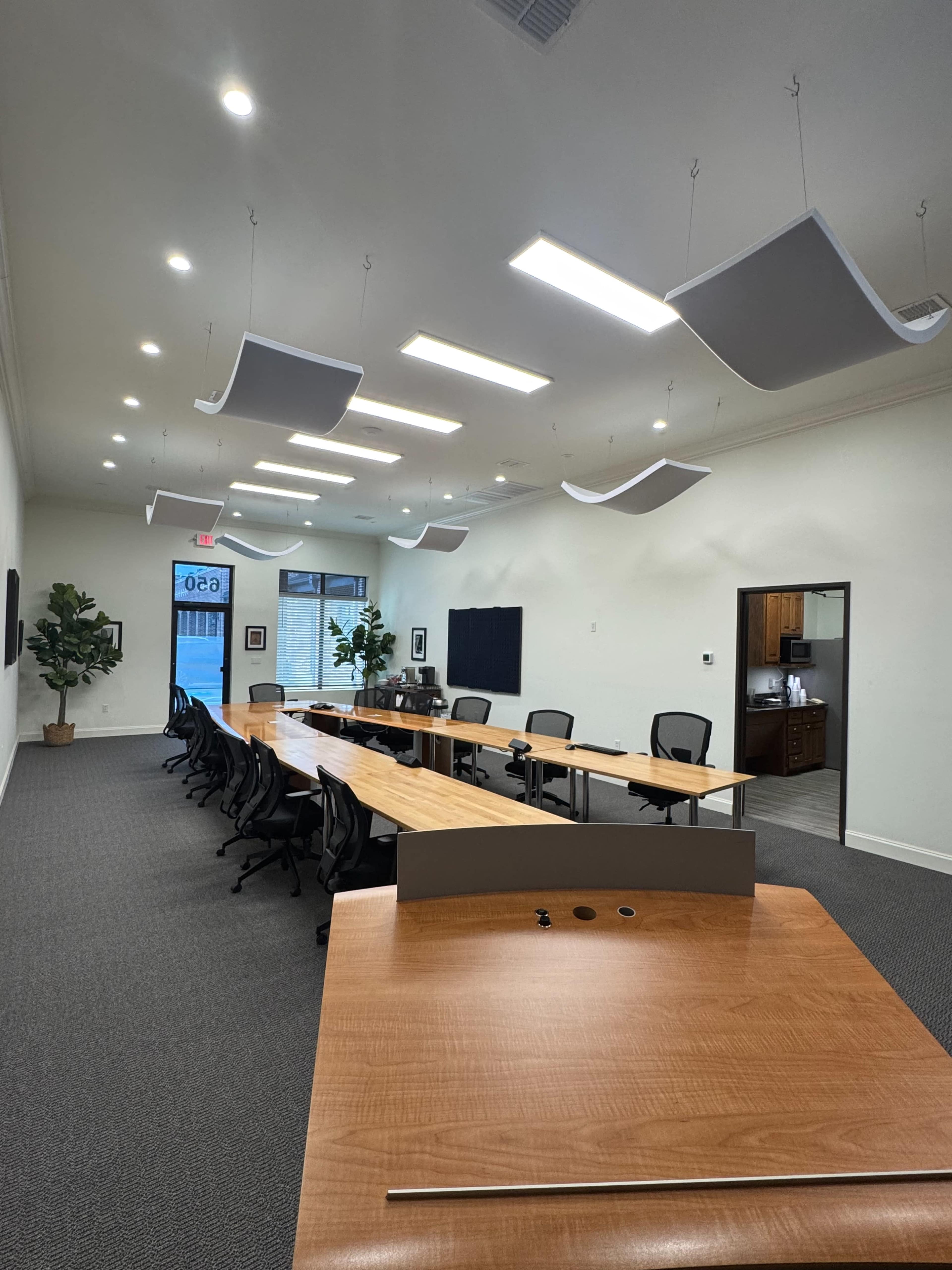 A modern conference room features a long wooden table surrounded by black chairs, with suspended acoustic panels above and a large screen mounted on the wall.