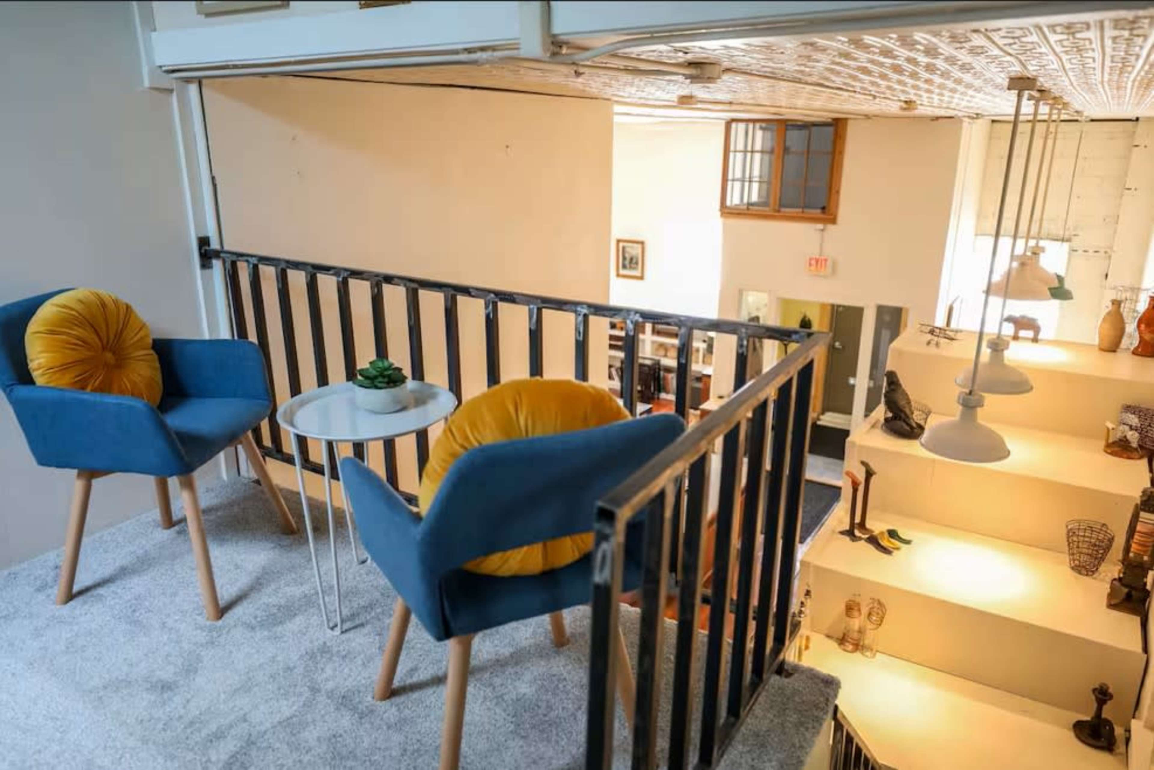 A small loft area features two blue chairs with yellow cushions and a side table overlooking a well-lit lower level embellished with decorative items on shelves.