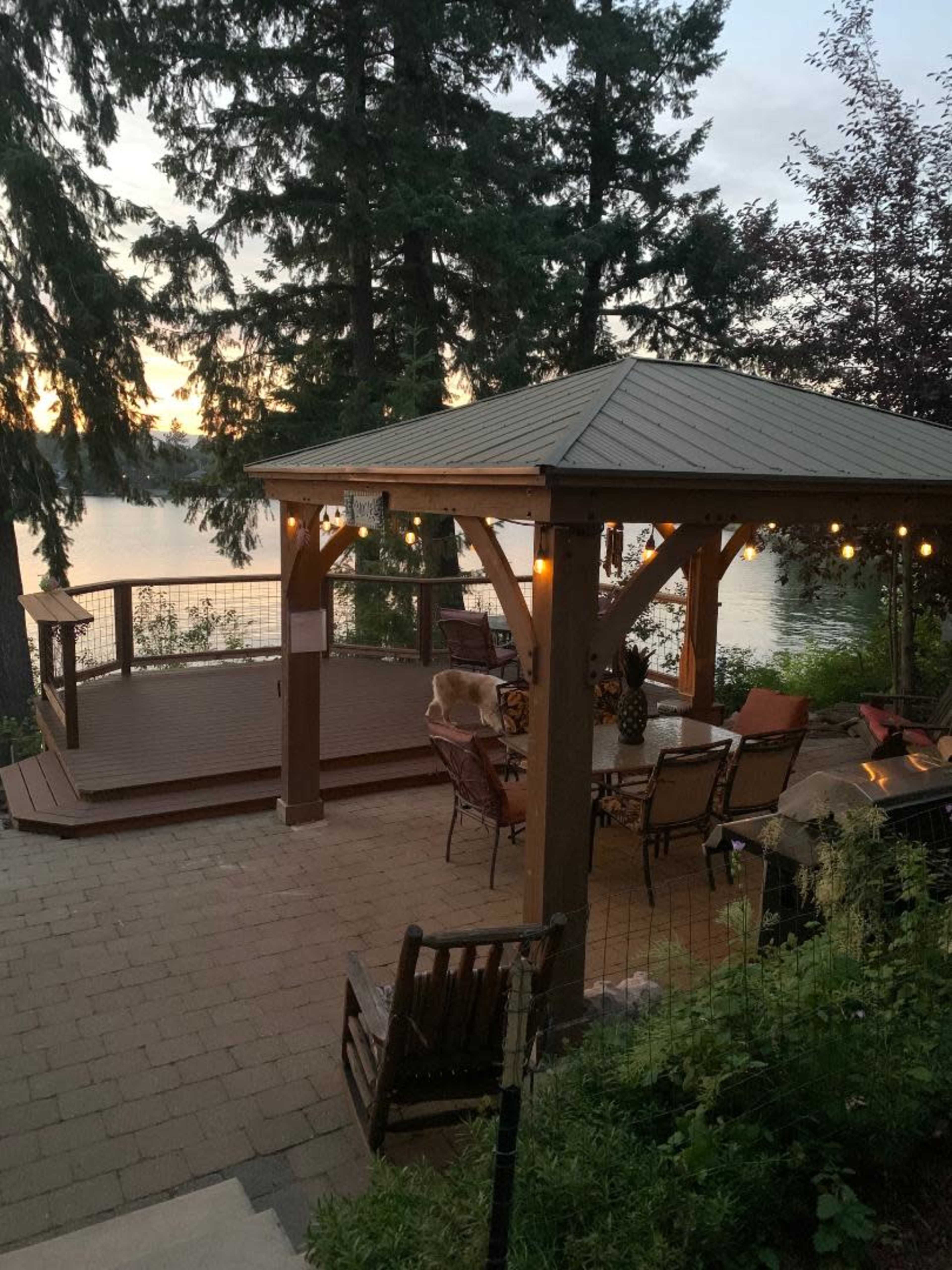 A wooden gazebo with string lights overlooks a patio area near a lake, surrounded by tall trees at sunset.