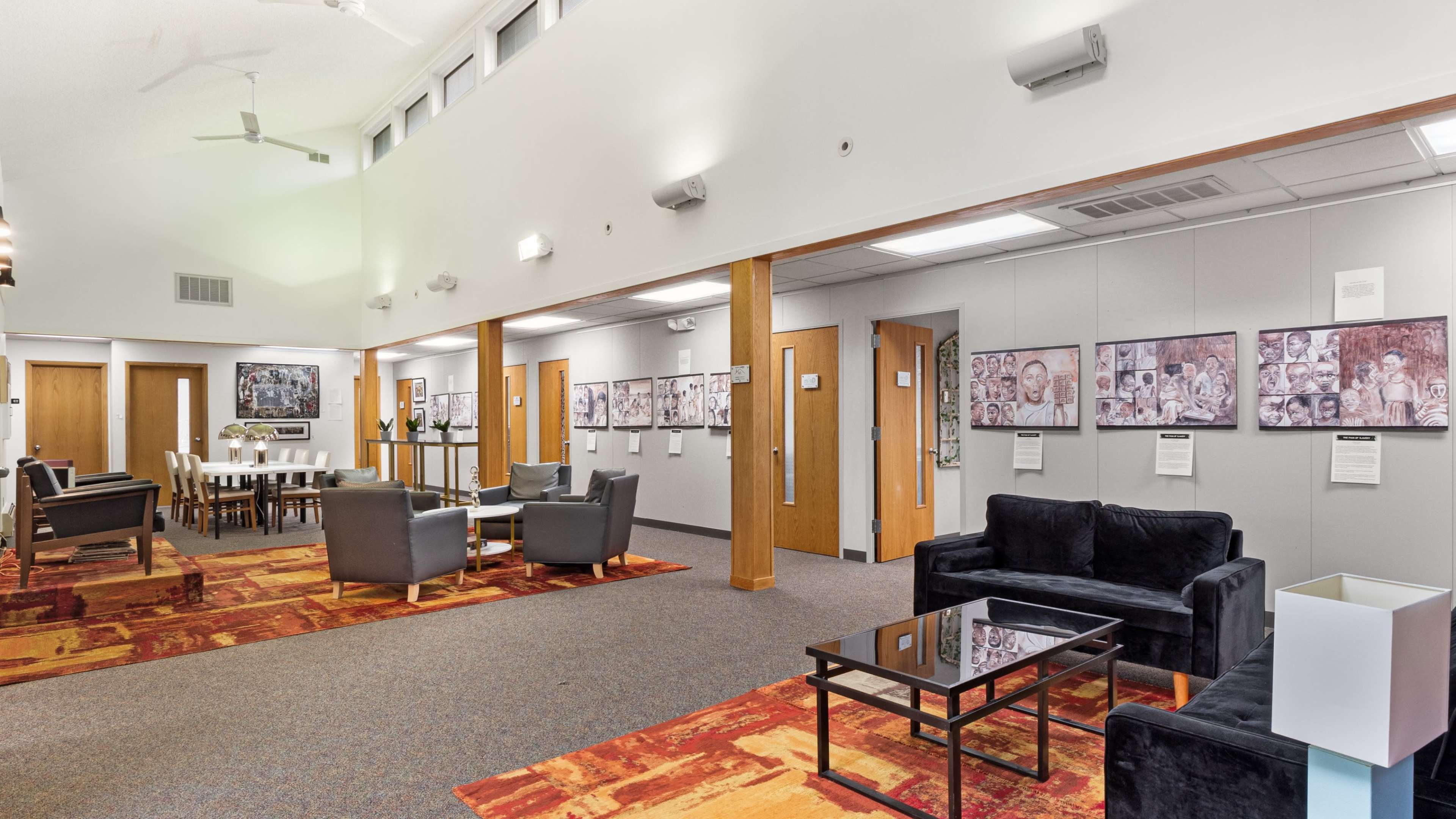 A spacious community room with a series of framed artworks on the walls, wooden doors, and seating areas arranged with black chairs and a glass coffee table.