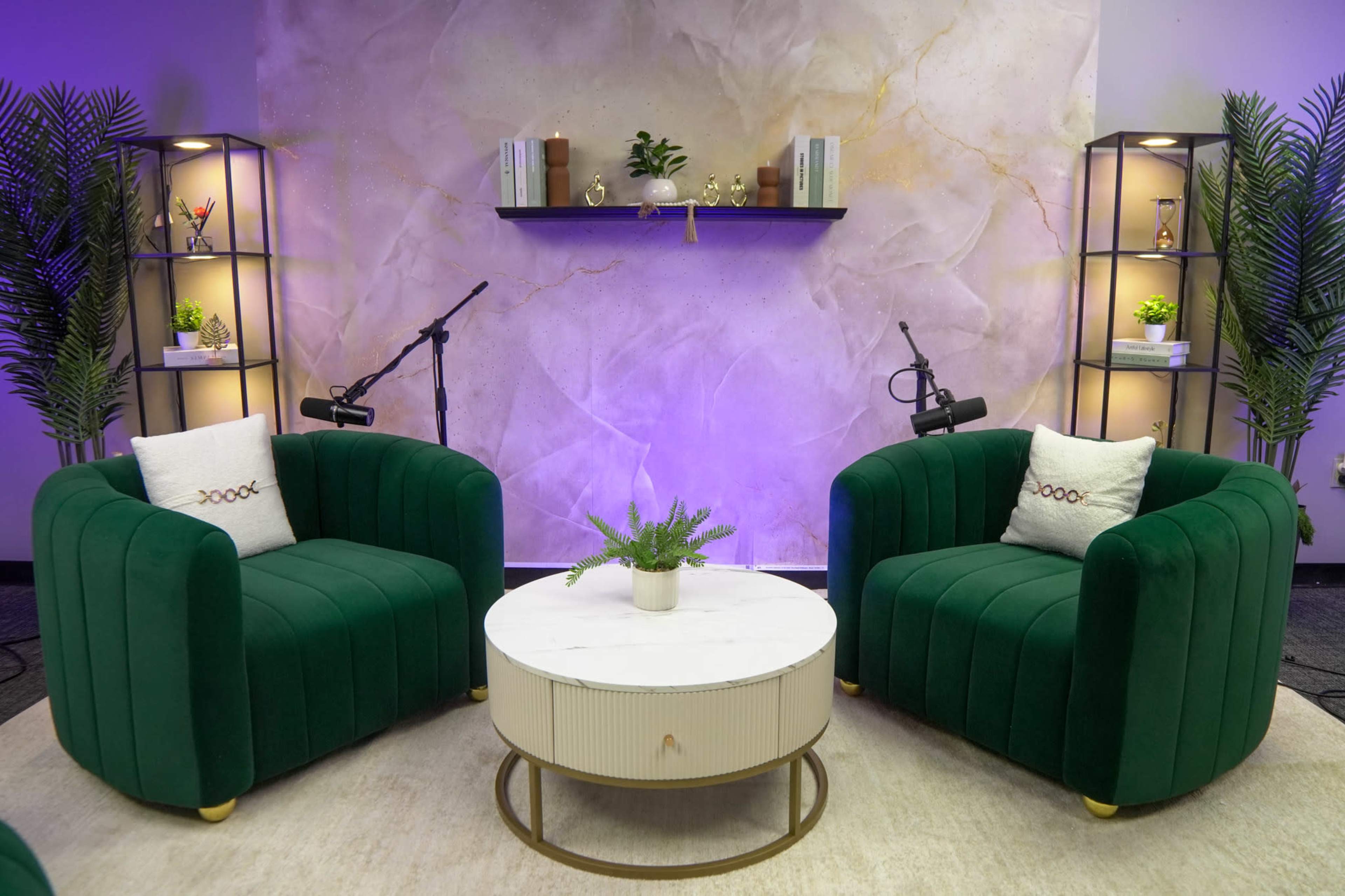 The image shows a cozy seating area with two green upholstered armchairs, a round marble coffee table, and shelf decorations against a softly illuminated, textured wall.