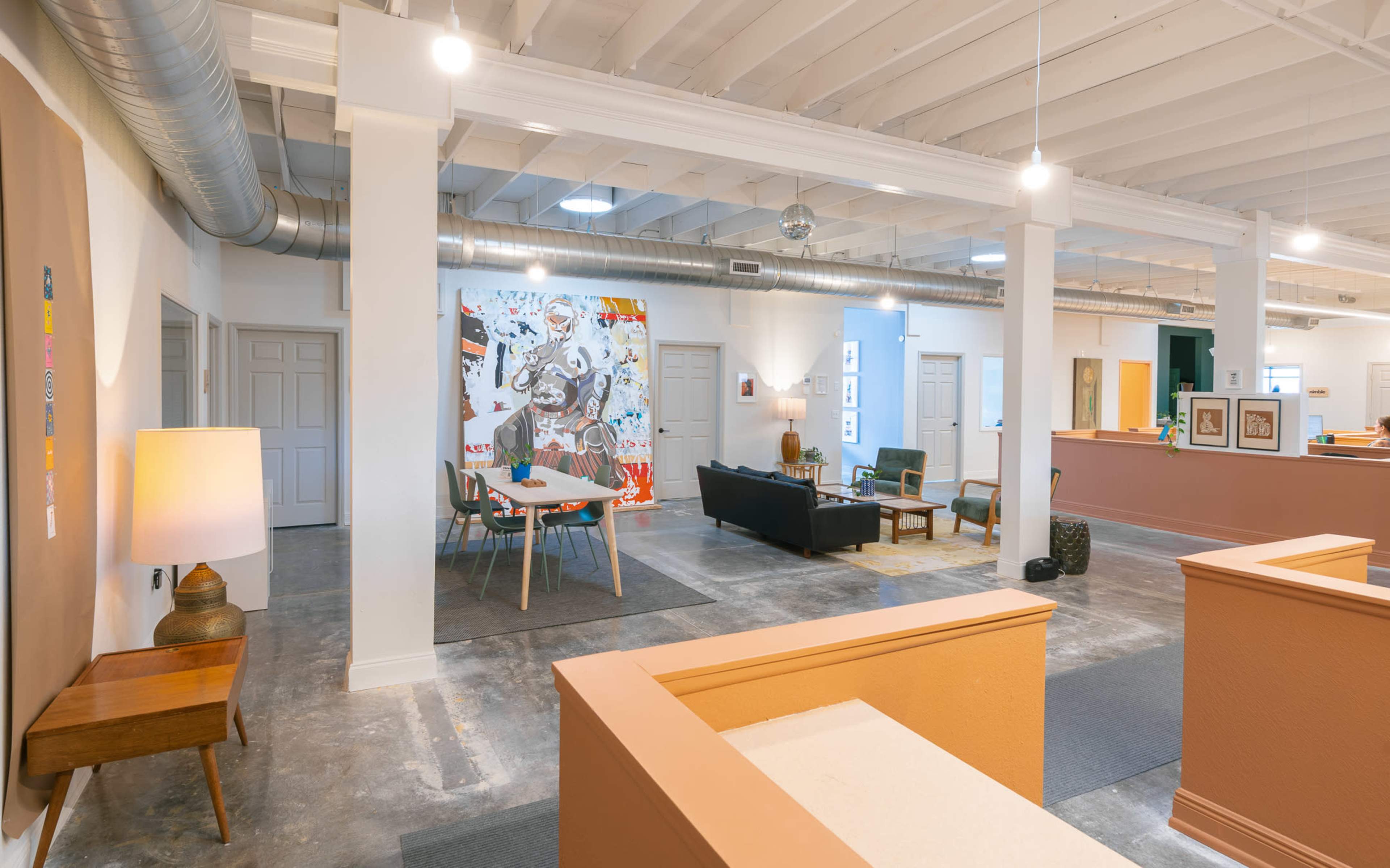 The image shows an open interior space featuring a seating area with a couch and chairs, a dining table, a large wall artwork, and light-colored walls with exposed ceiling beams.