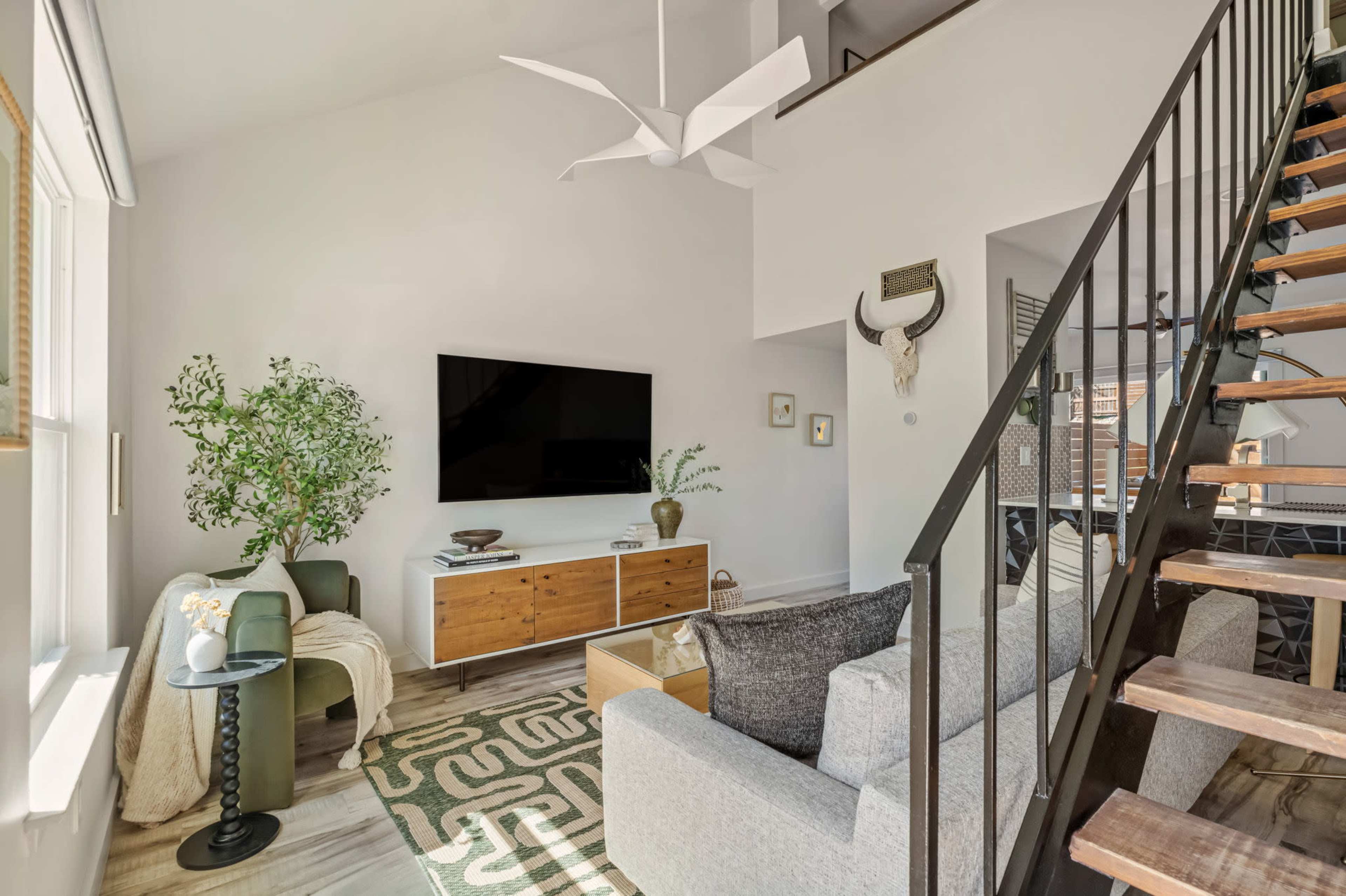 The image shows a modern living room with a gray sofa, wooden media console, large flat-screen TV, an indoor plant, and a staircase leading to an upper level.