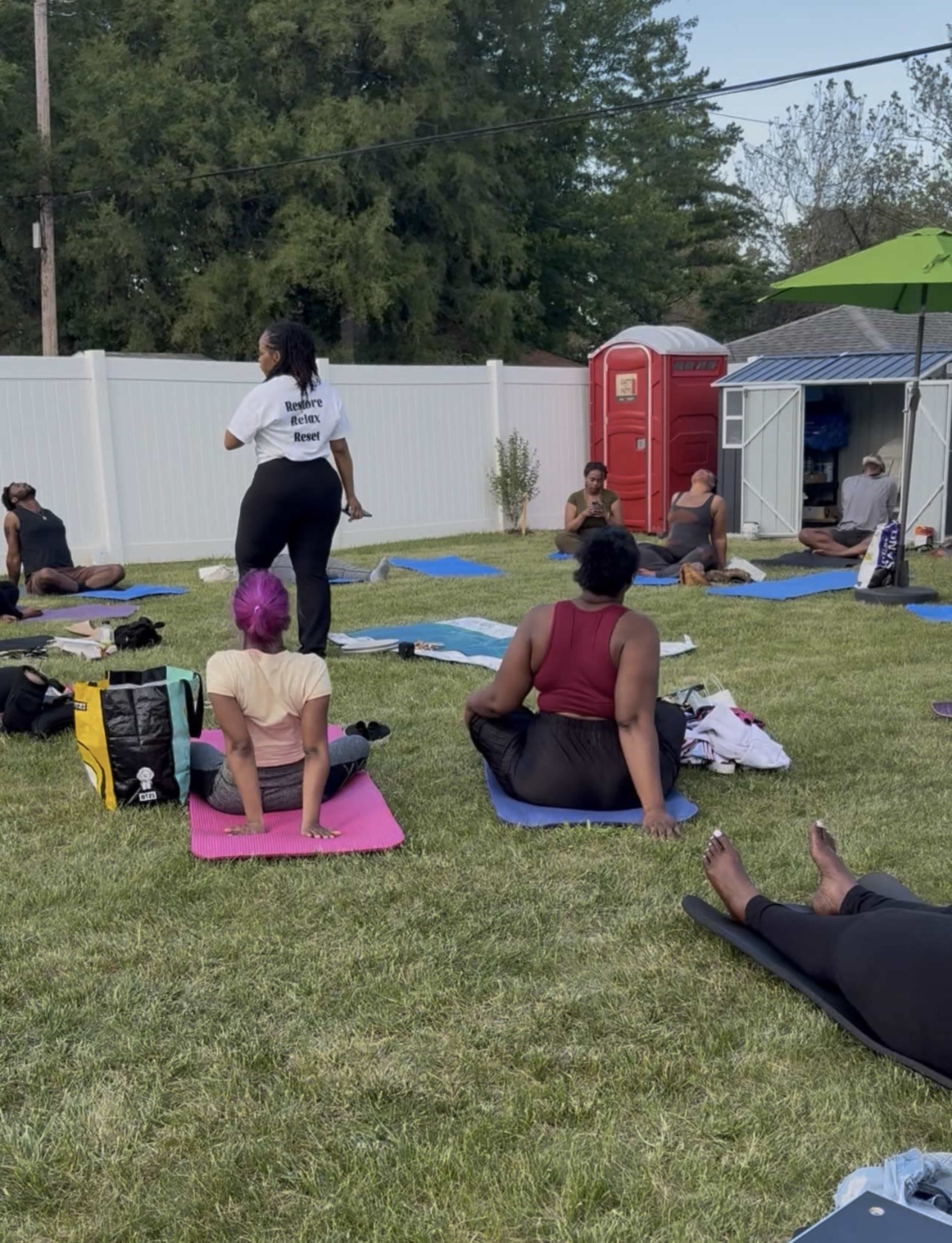A group of people are practicing yoga on mats in a grassy outdoor area, while an instructor stands in front of them.