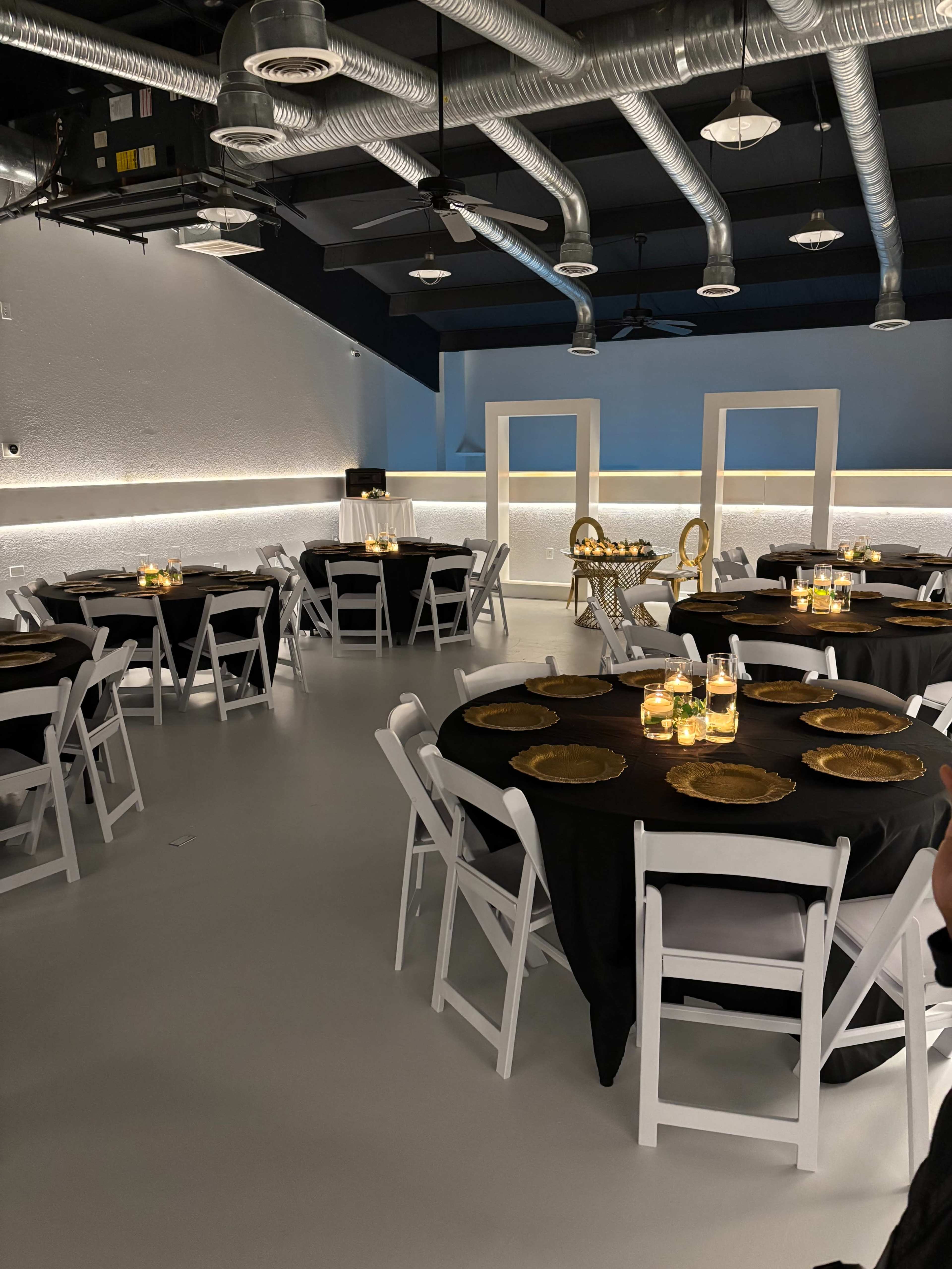 The image shows a spacious venue set up for an event, featuring round tables with black tablecloths, gold chargers, and candles, surrounded by white chairs.