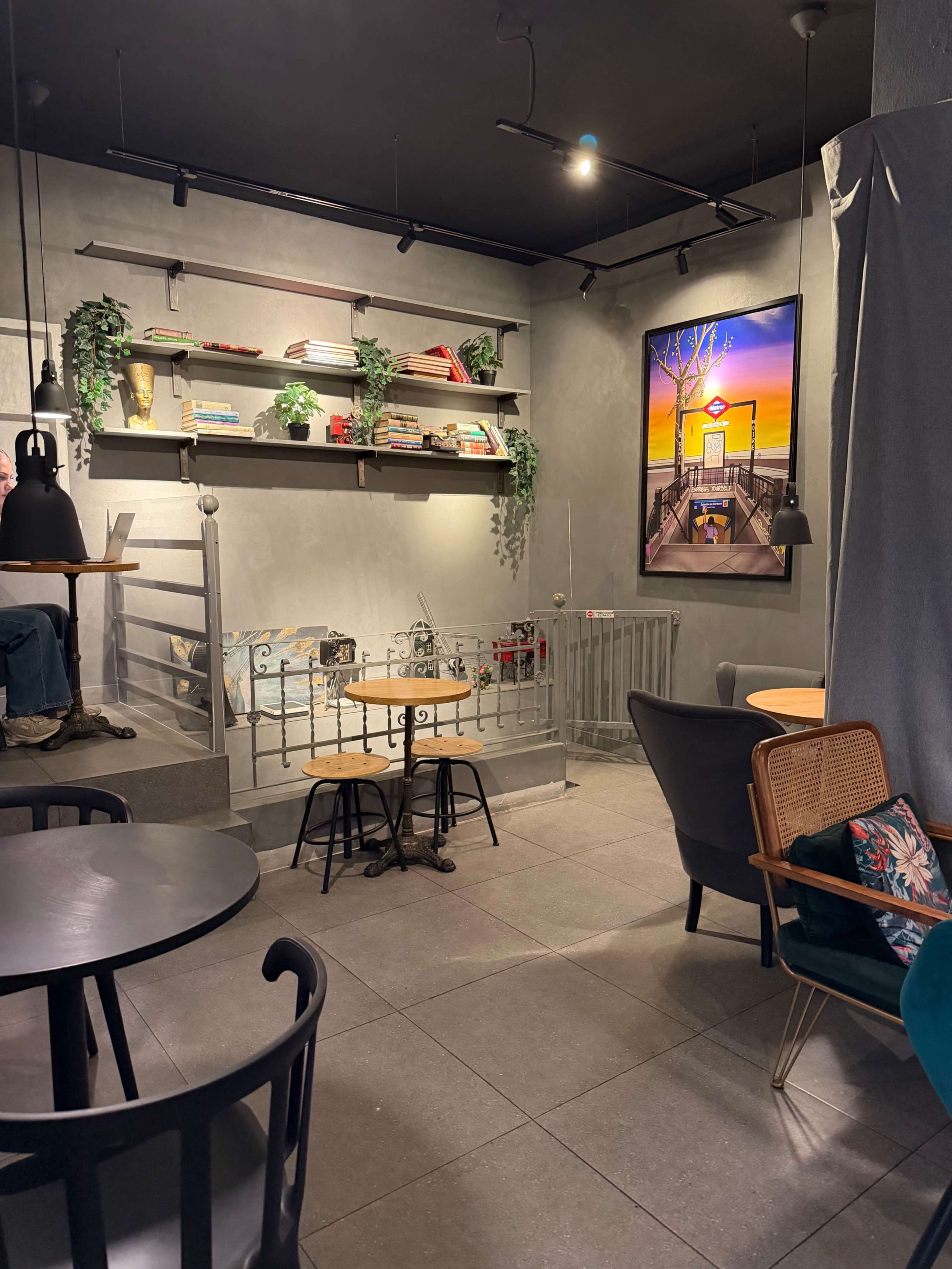 The image shows a cozy cafe interior featuring wooden tables, a shelf filled with books and decorative items, and a wall-mounted artwork depicting a sunset scene.