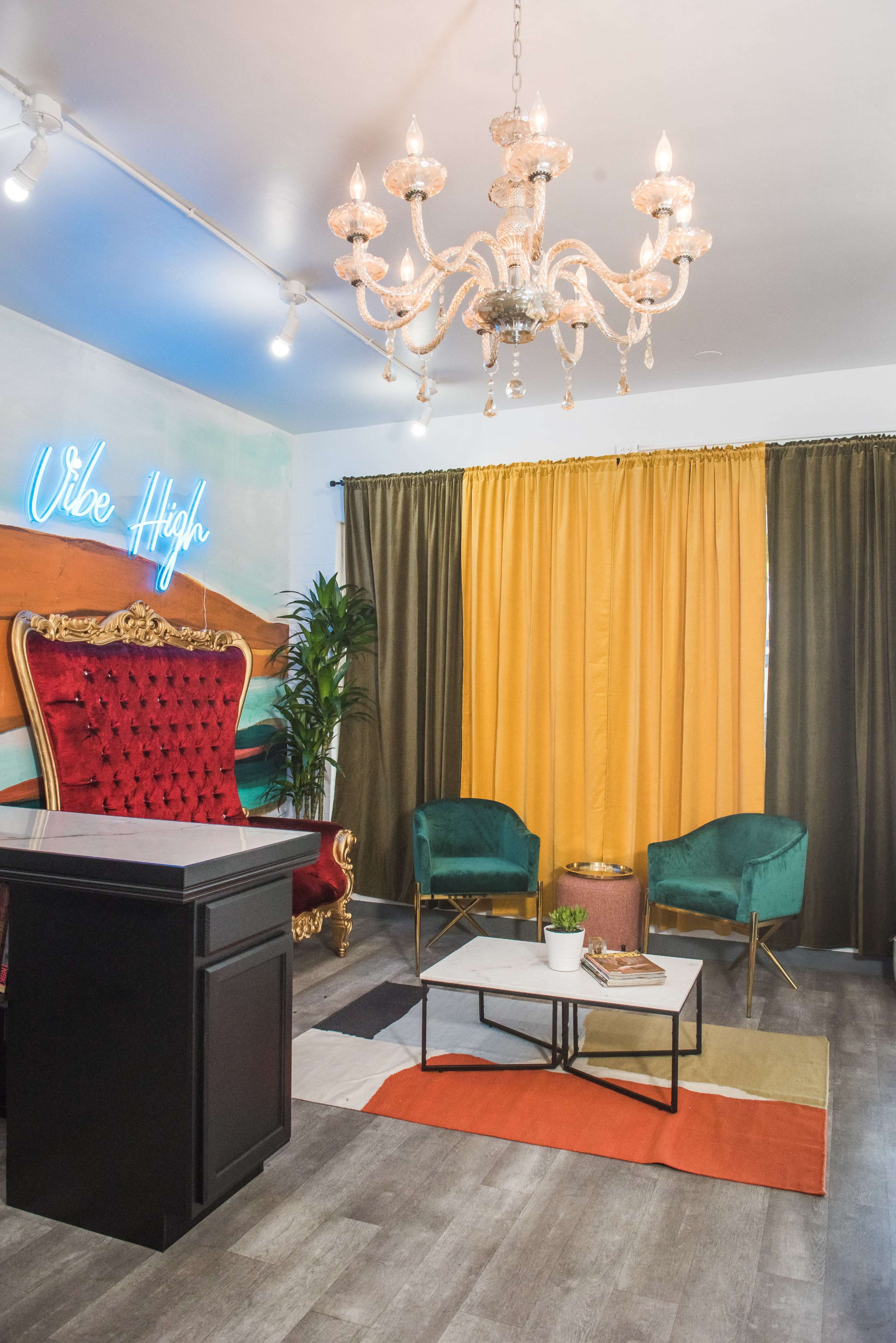 The image shows a stylish room featuring a large red throne chair, a chandelier, two green chairs, a dark reception desk, and colorful curtains.