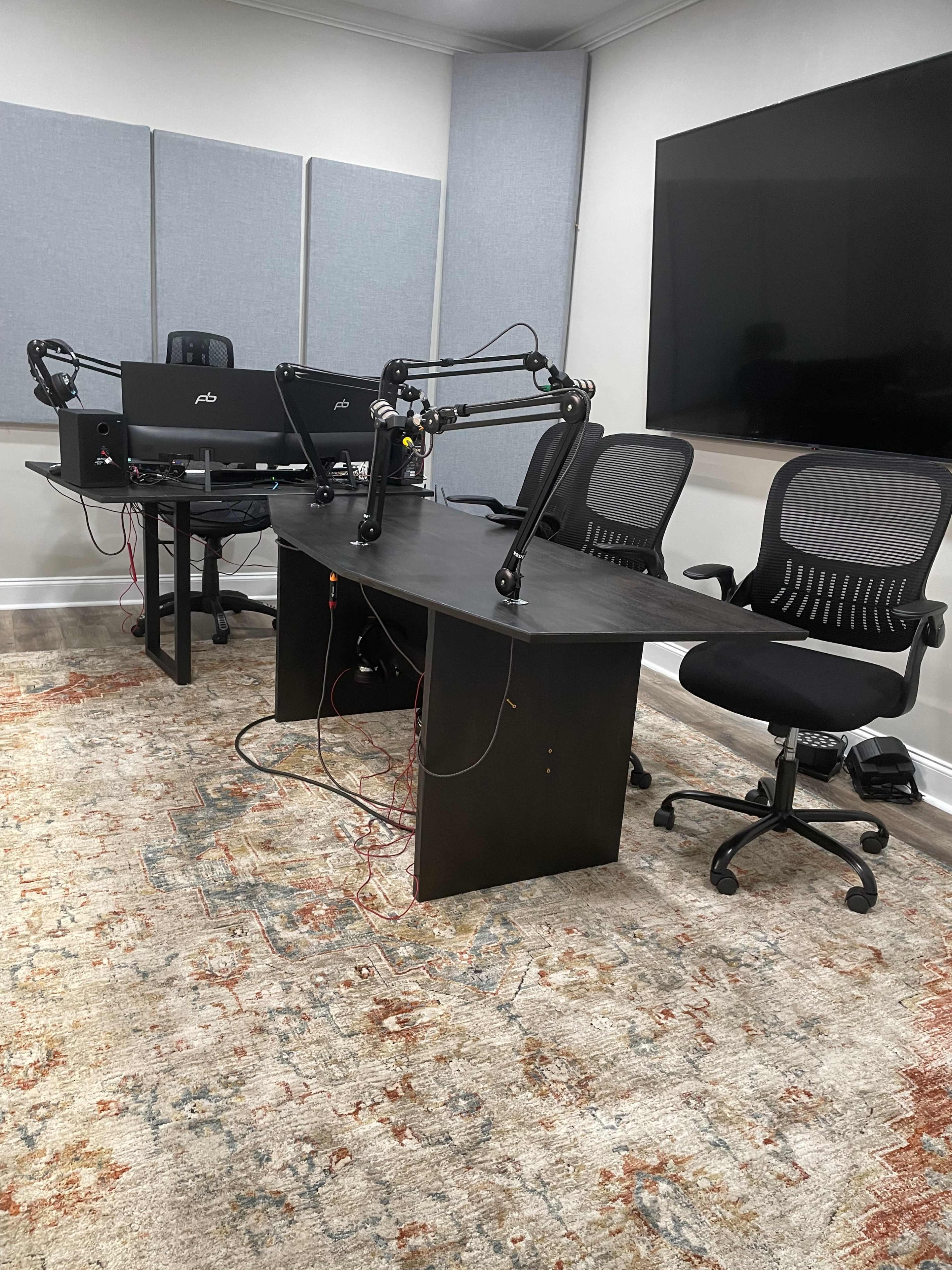 The image shows a modern recording studio setup with a large table, three chairs, multiple microphones, and two computer monitors.