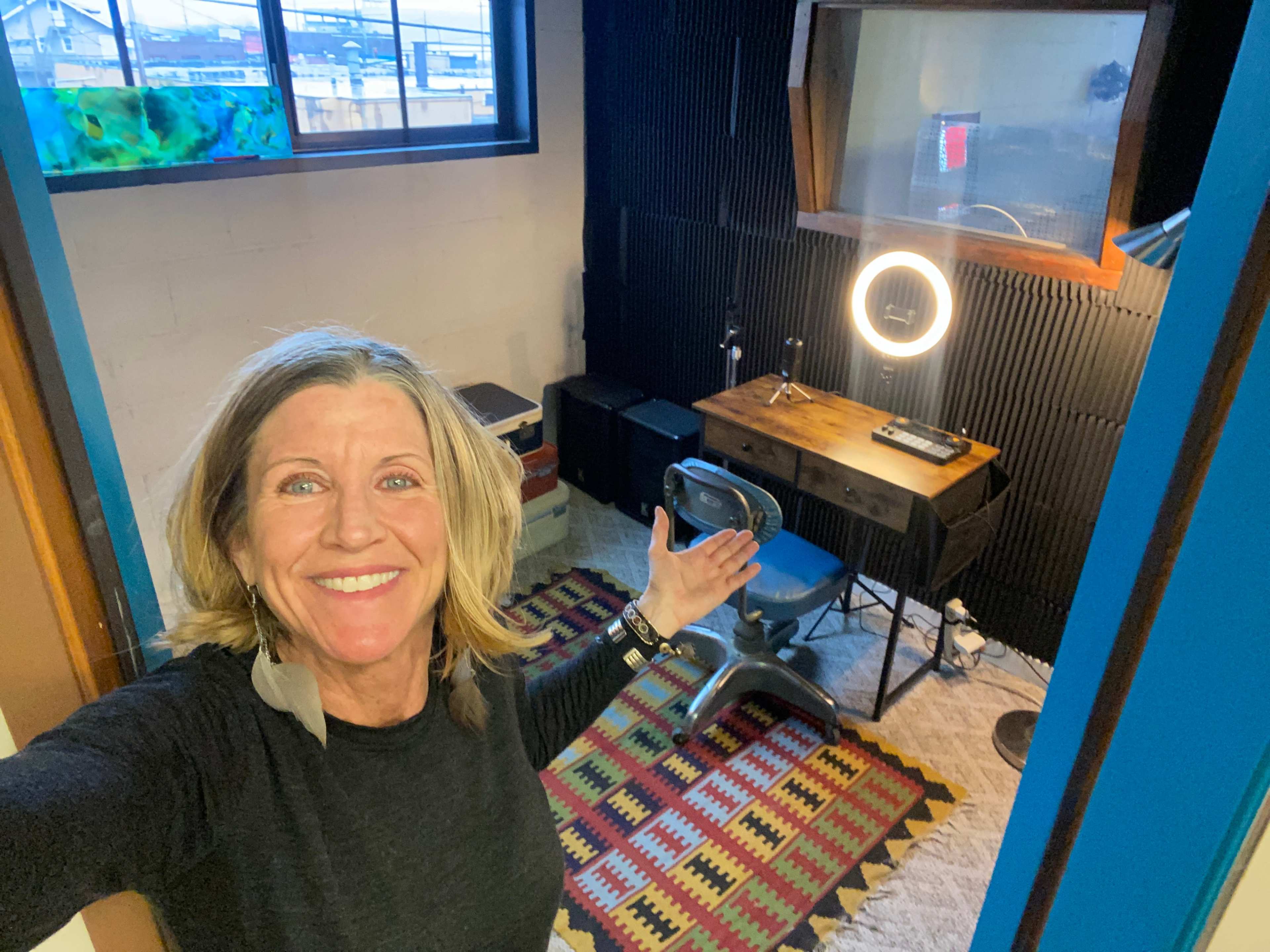 A woman stands in a small room that features a desk with a lamp, a chair, and a patterned rug.
