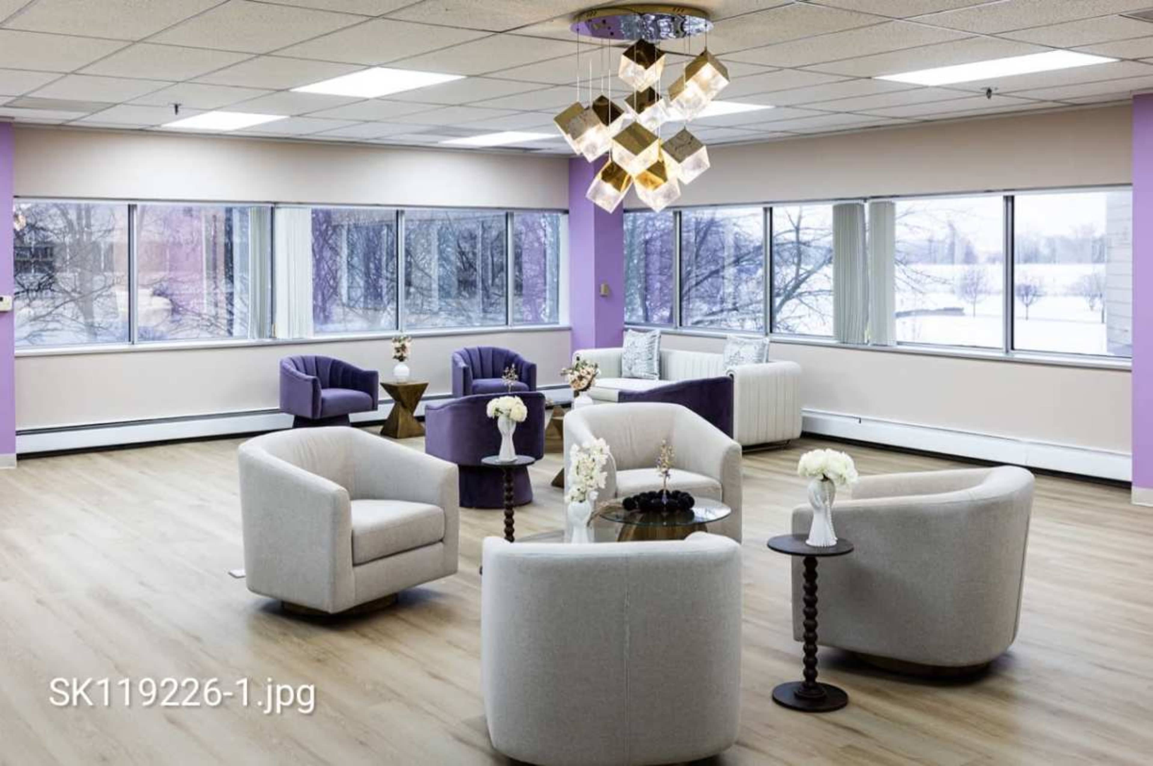 The image depicts a modern, well-lit lounge area featuring several chairs and a central table, with large windows showing a view of the outdoors.