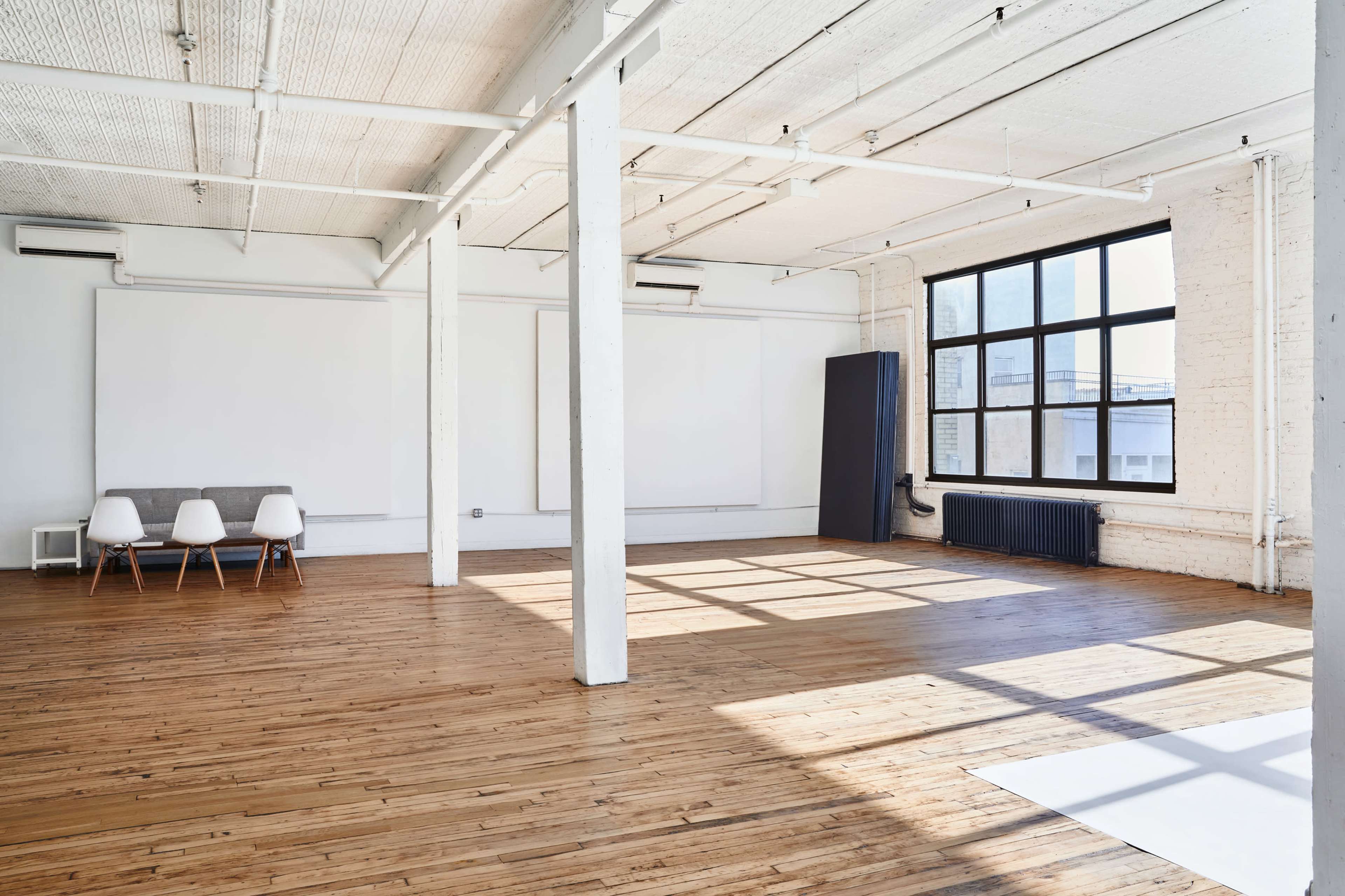 A spacious, well-lit room with wooden floors, large windows, and a few white chairs arranged near the wall.