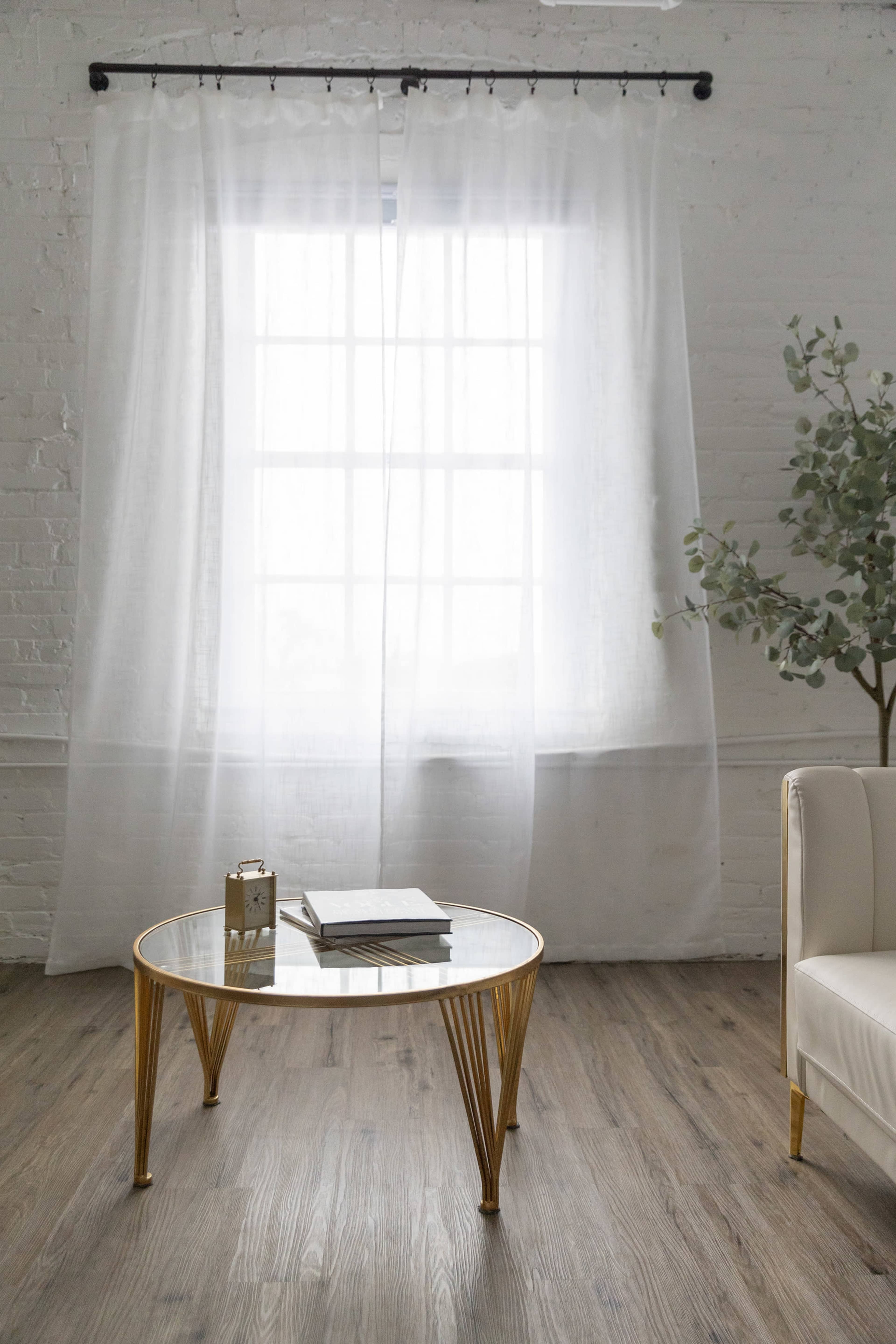 A bright room features a round glass coffee table with gold legs, a stack of books, and a small decorative item, alongside a white sofa and sheer curtains draping over a large window.
