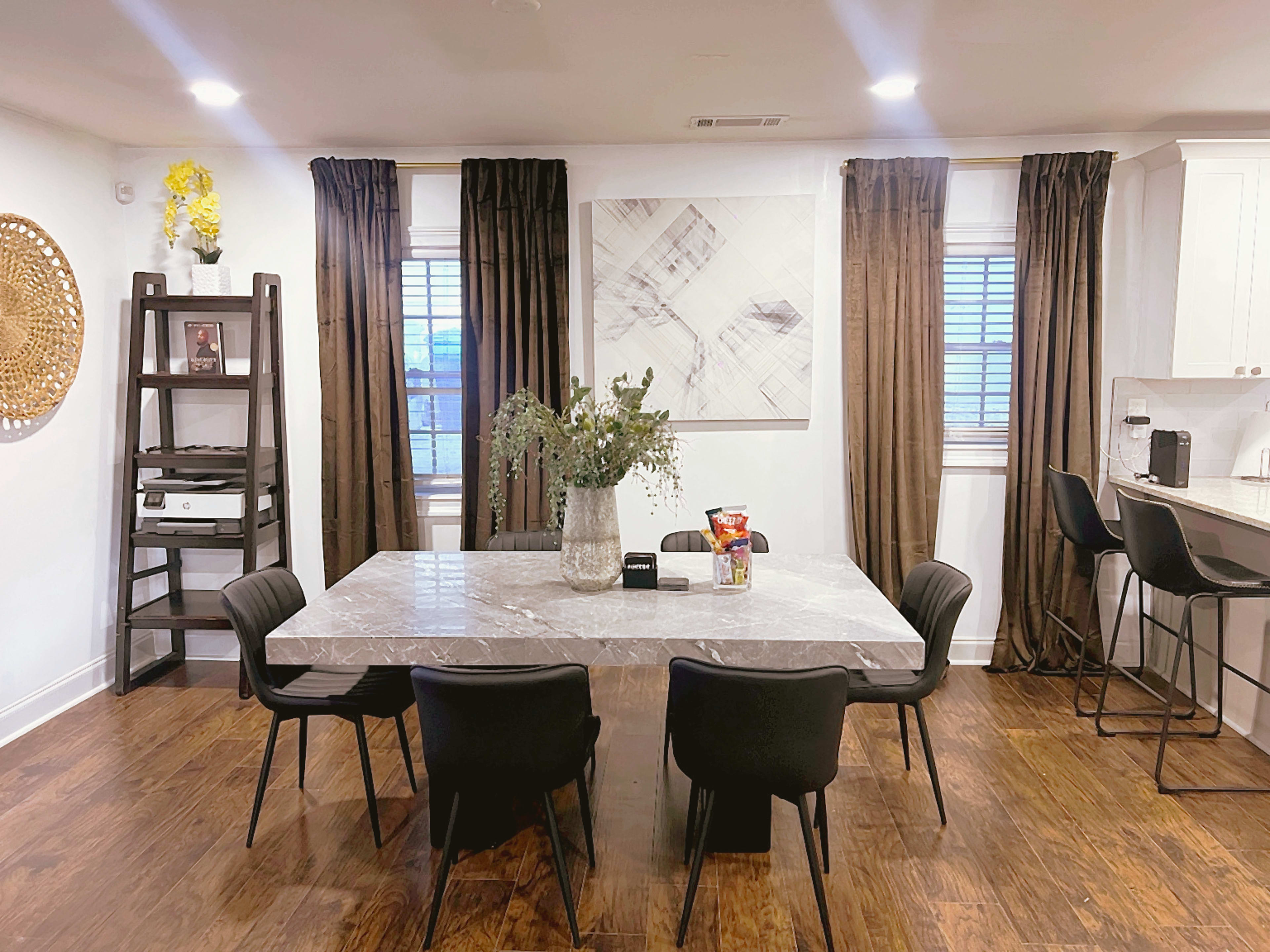 The image shows a dining area with a marble table surrounded by black chairs, a large wall art piece, and dark curtains framing the windows.