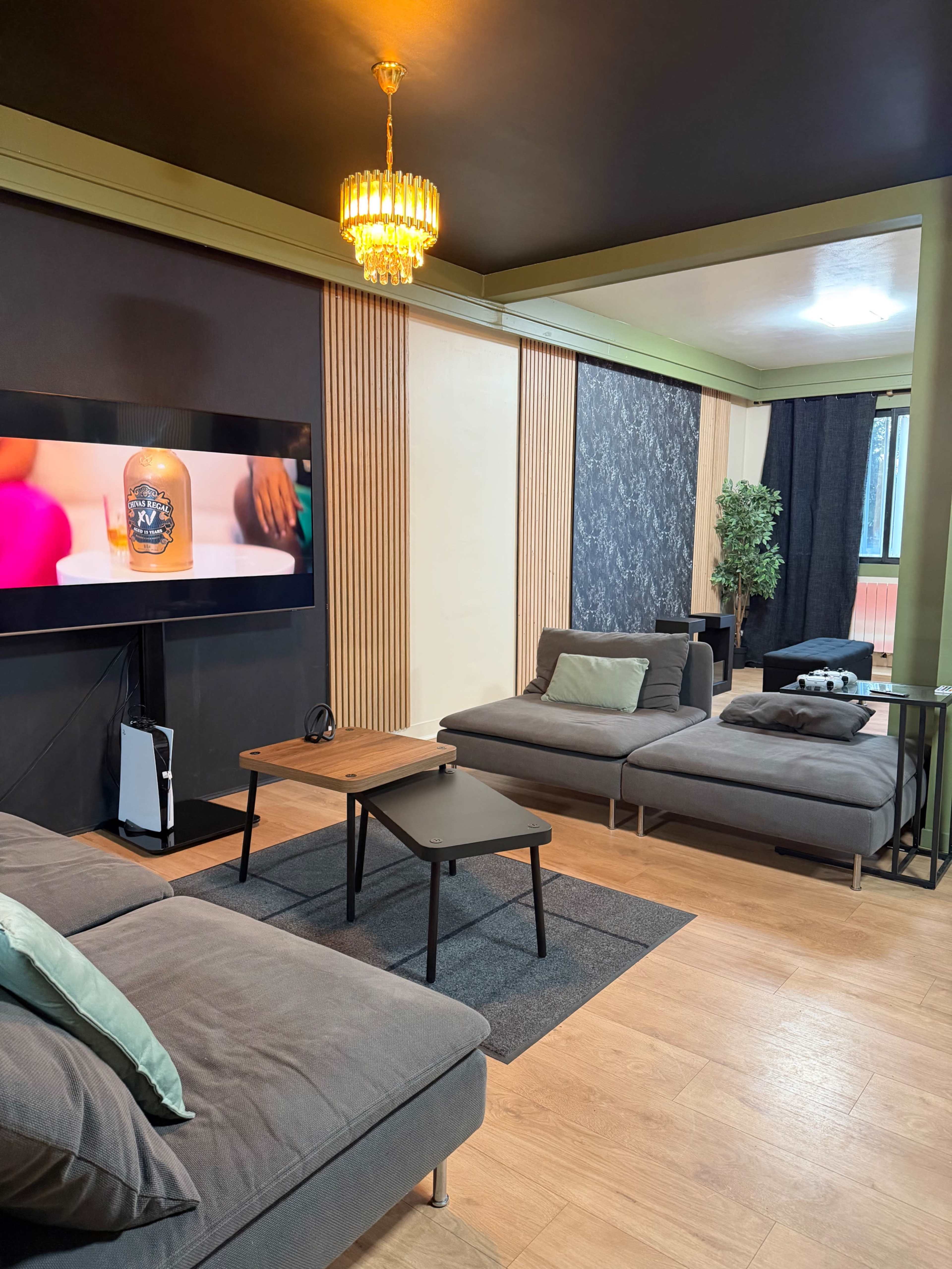 The image shows a modern living room featuring gray sofas, a coffee table, a flat-screen TV on the wall, and a decorative plant in the corner.