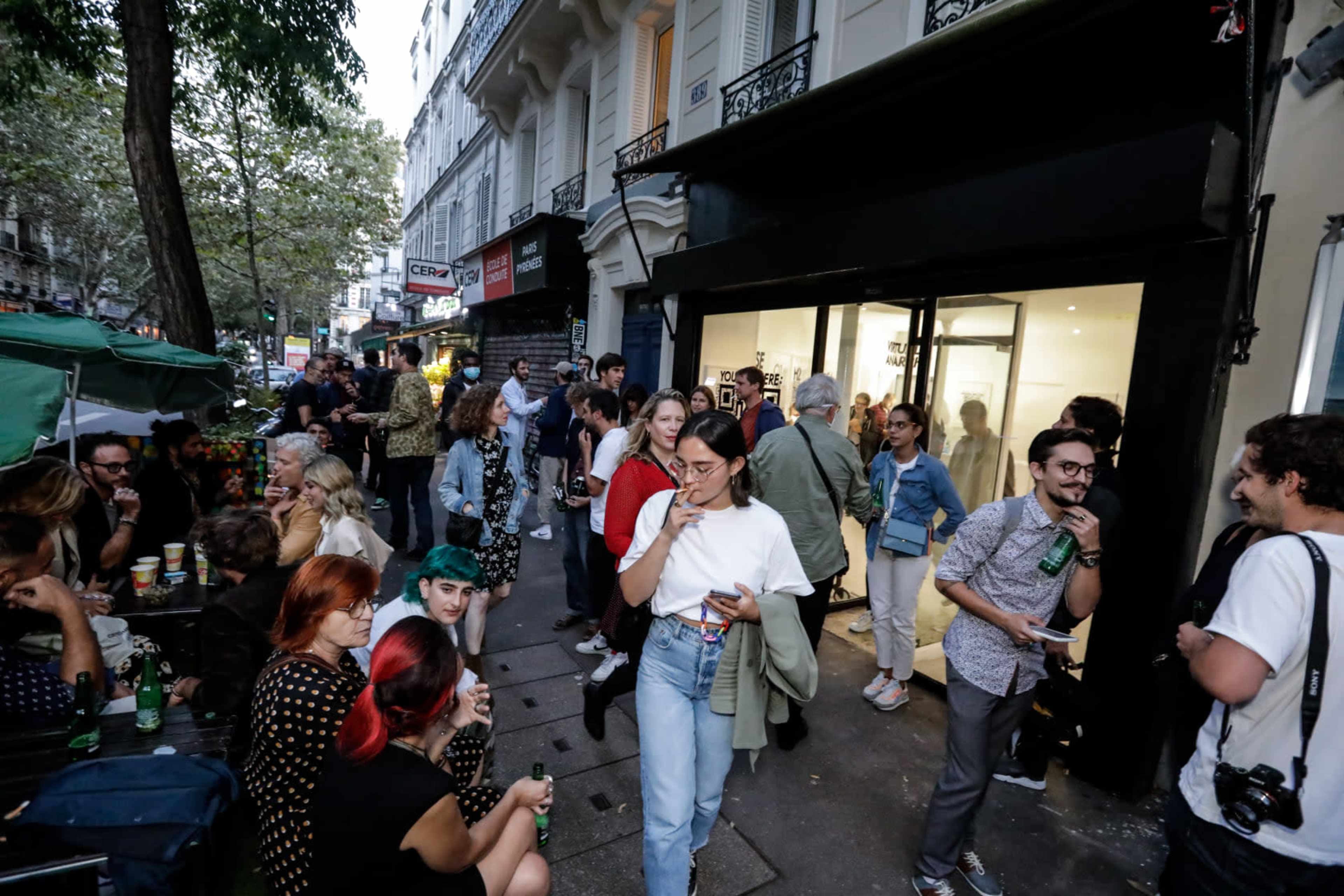 A crowd gathers outside a storefront on a bustling street, with people socializing and holding drinks.
