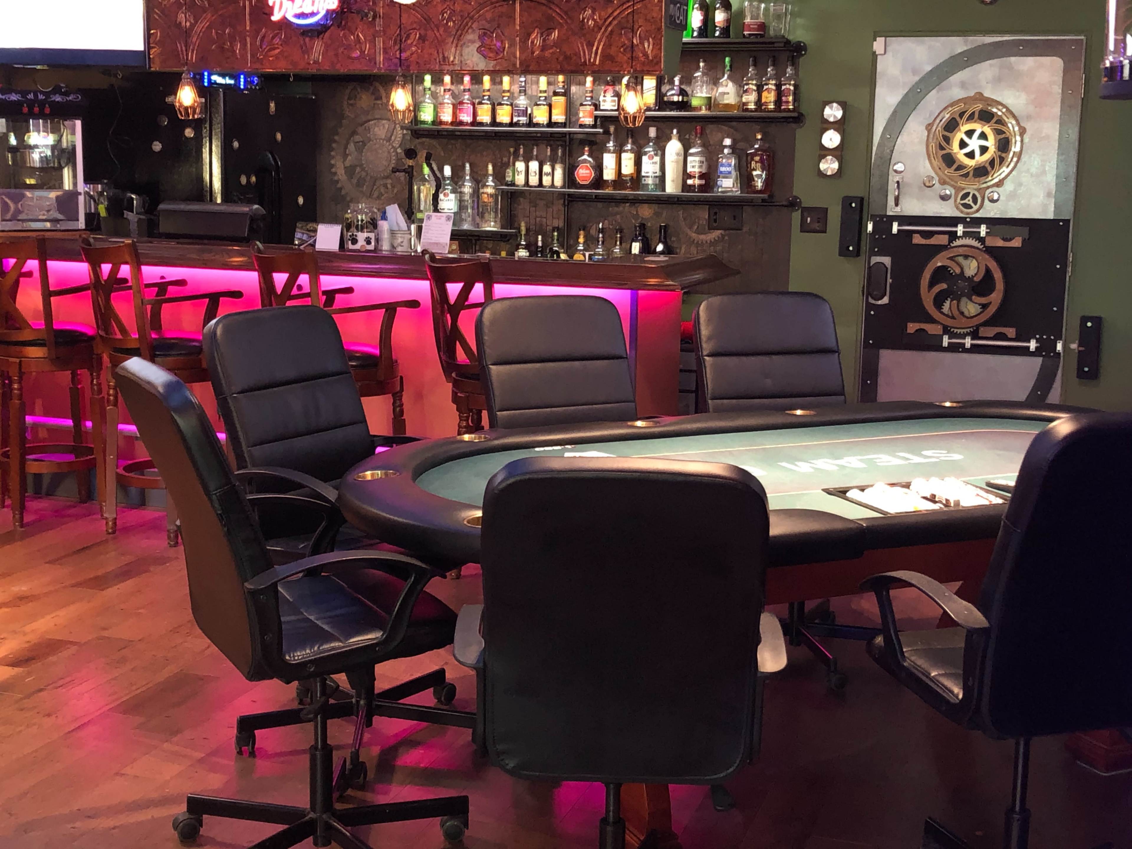 A poker table surrounded by several black chairs in a dimly lit bar setting with a backlit shelf of liquor bottles.