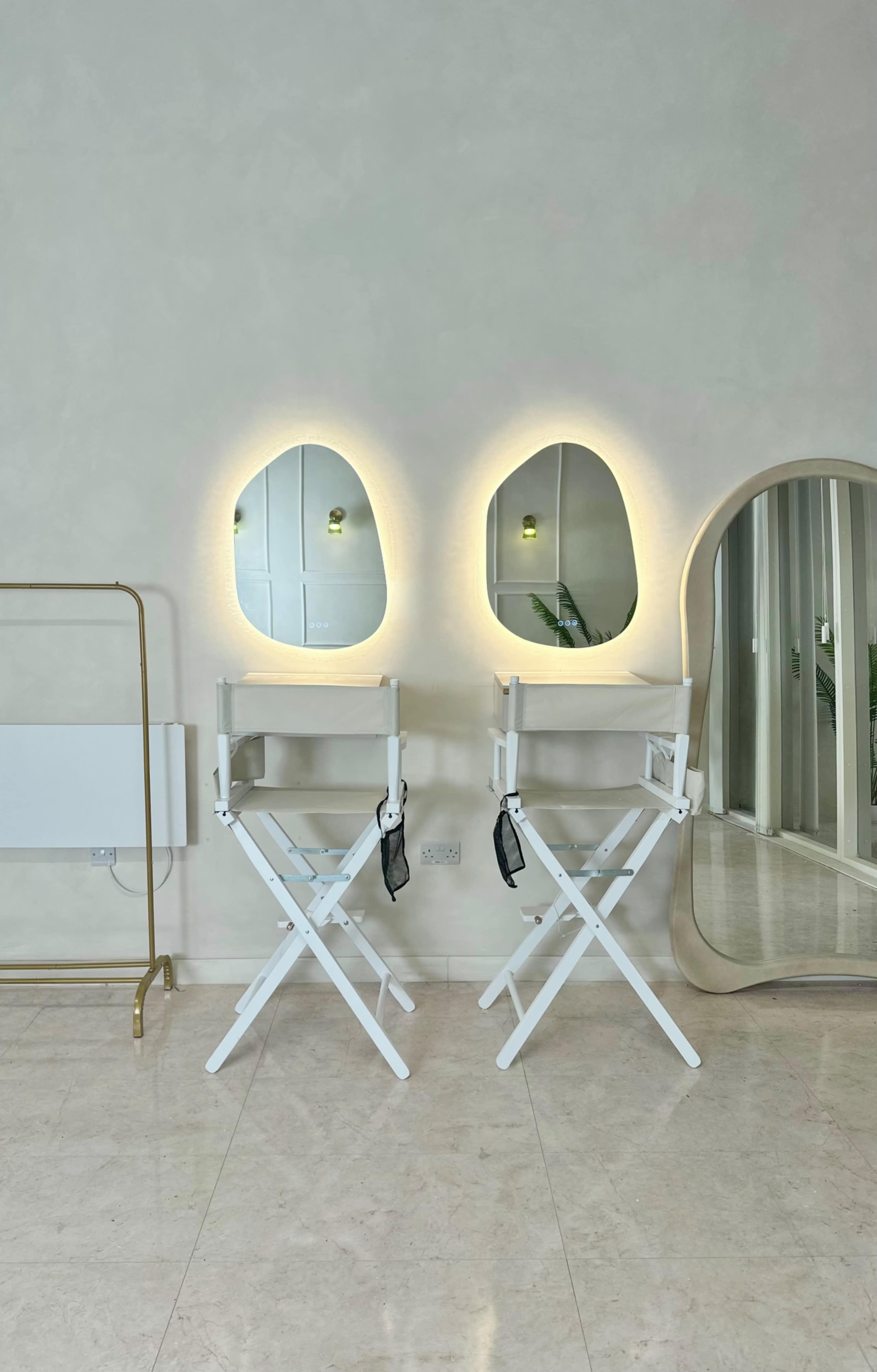 The image shows two director's chairs facing mirrors with soft, rounded edges, set against a light-colored wall in a spacious room.
