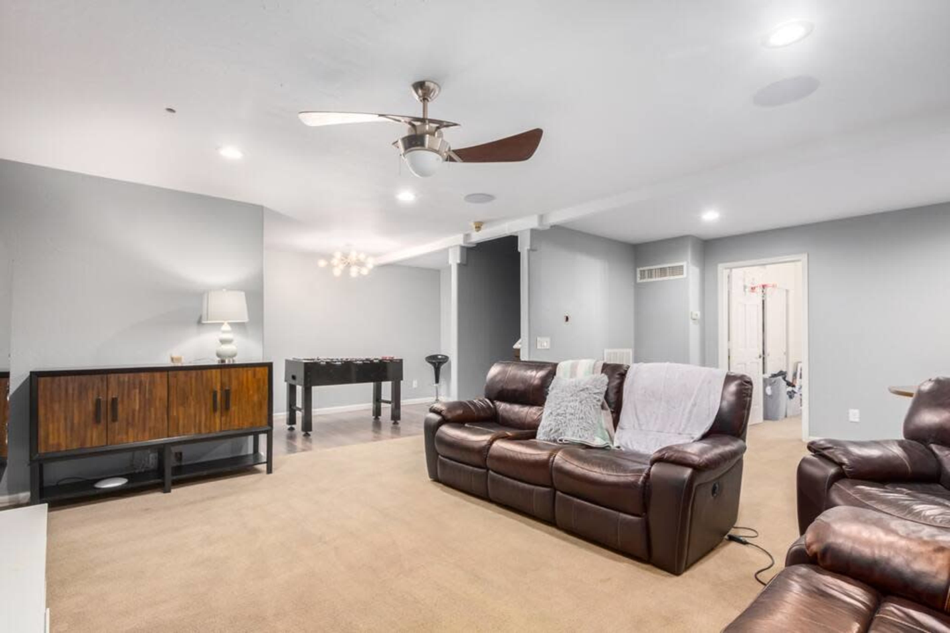A spacious living room with brown leather sofas, a light-colored carpet, a ceiling fan, and a foosball table.